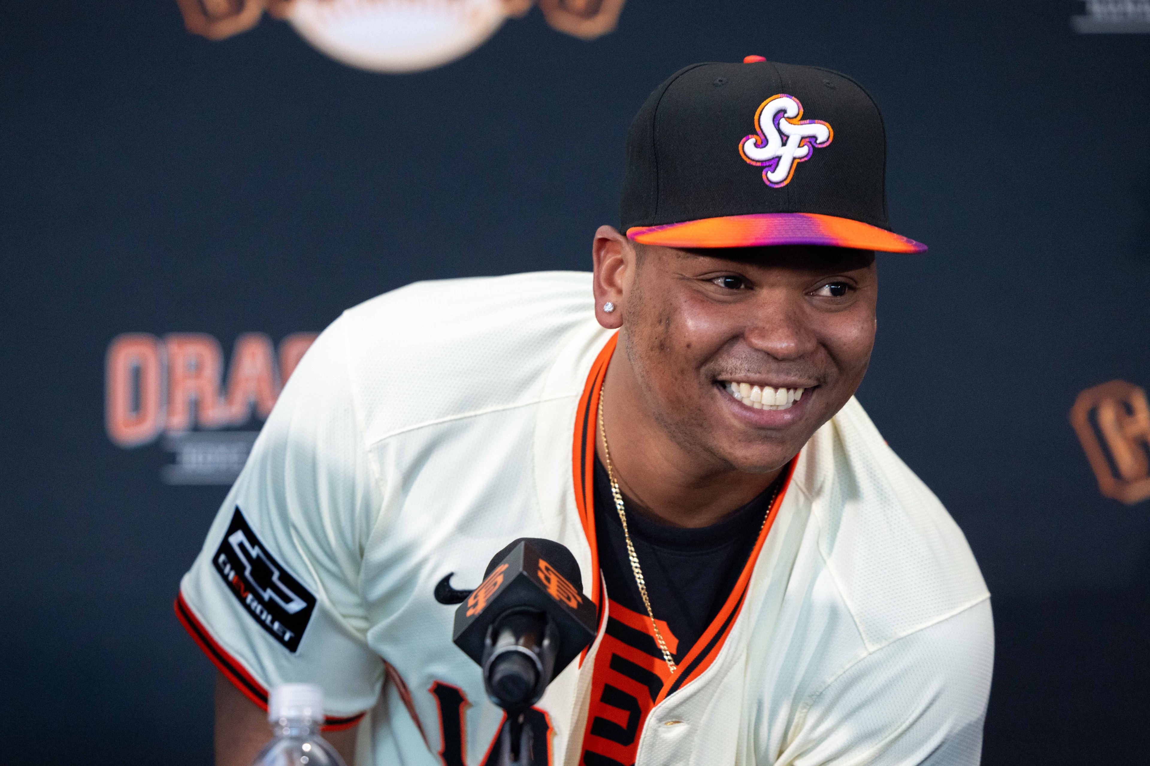 A smiling man wearing a black cap with "SF" on it and a white baseball jersey leans towards a microphone at a press event, with logos behind him.