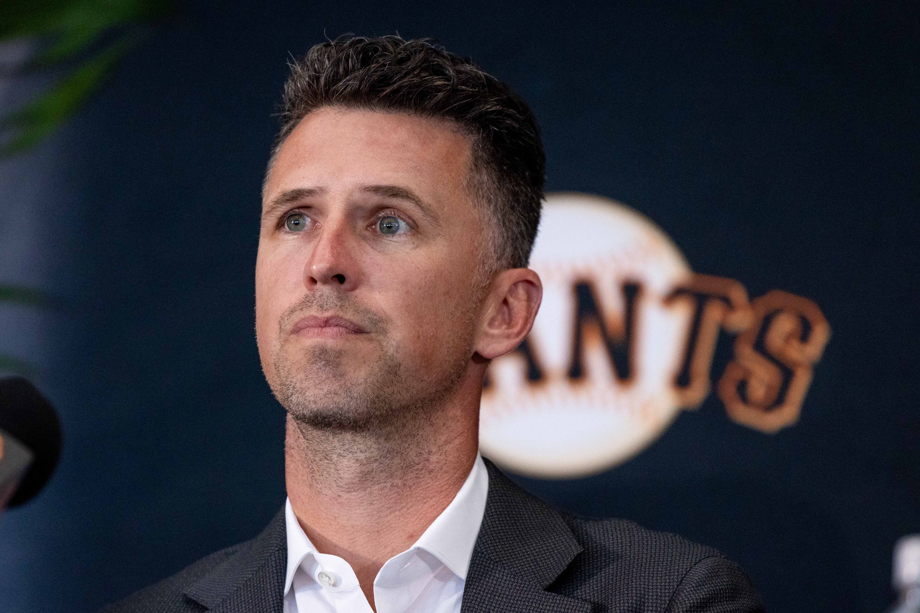 A person with short hair is wearing a suit and looking thoughtful. Behind them is a wall with a design resembling a baseball and the word "GIANTS."