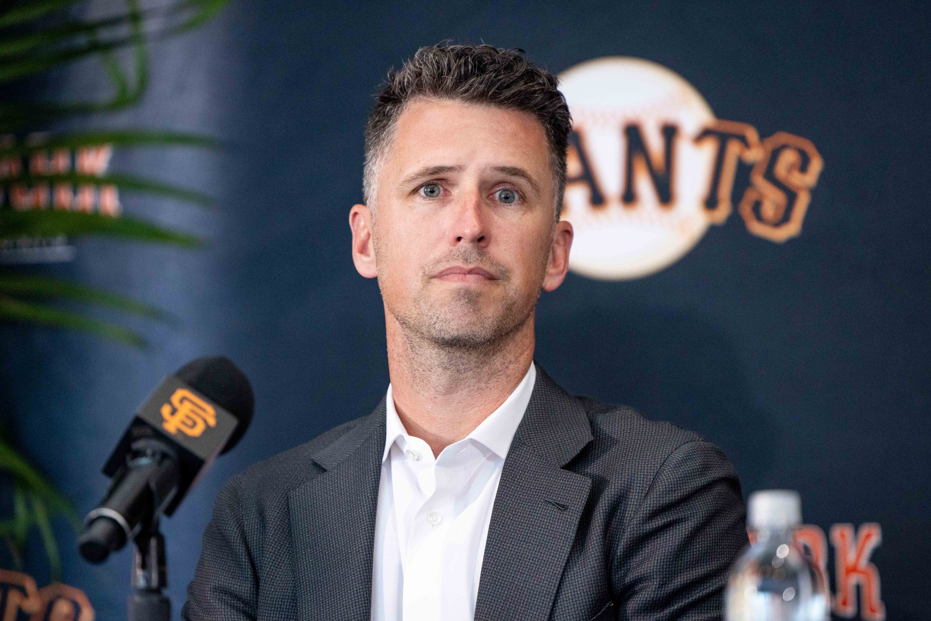 A person in a suit sits at a press conference with a mic displaying the San Francisco Giants logo. The backdrop shows the team's name partially visible.