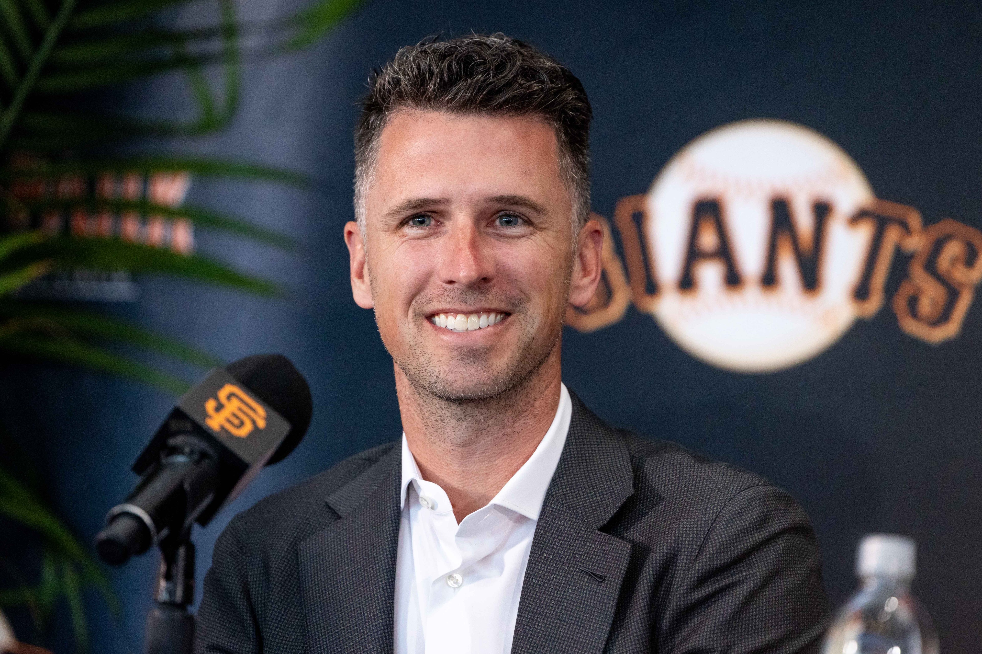 A person in a suit smiles at a press conference with a microphone featuring a logo. A "Giants" logo on a baseball is visible in the background.