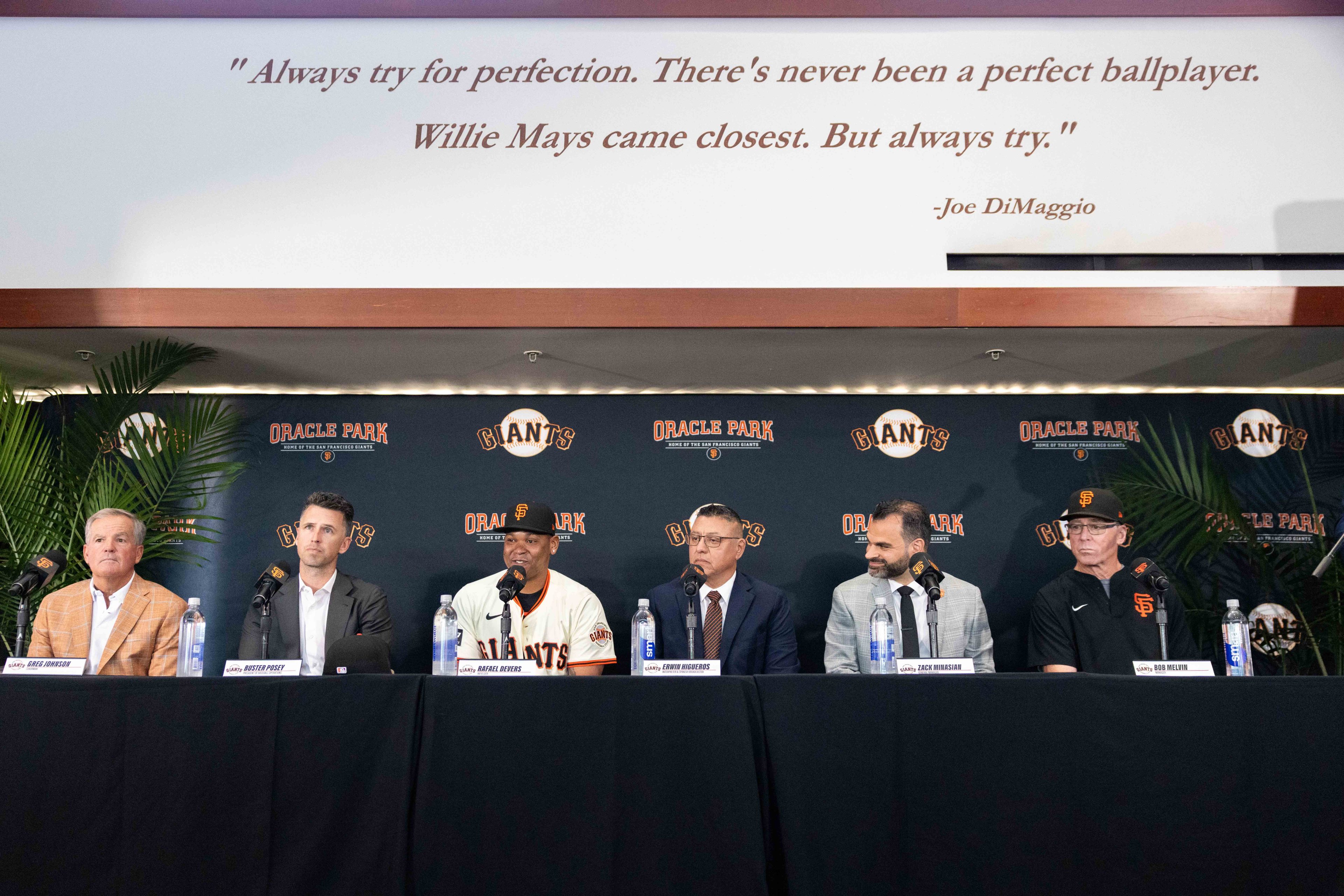 Six men sit at a table with microphones in front of a Giants backdrop, under a quote by Joe DiMaggio. Some are in suits, and one wears a Giants jersey.