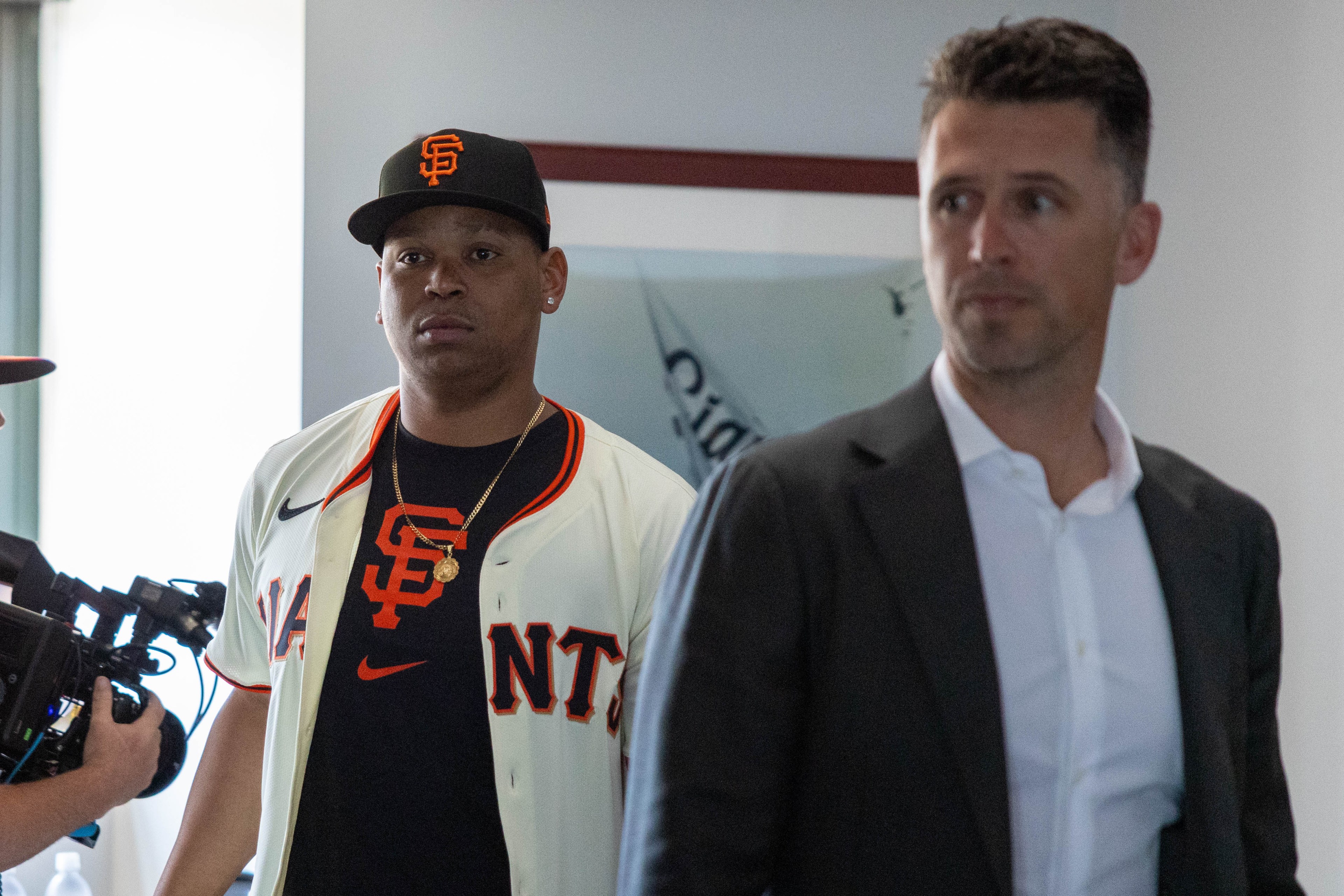 A man in a baseball jersey and cap stands beside another man in a suit. A camera operator is partially visible on the left.