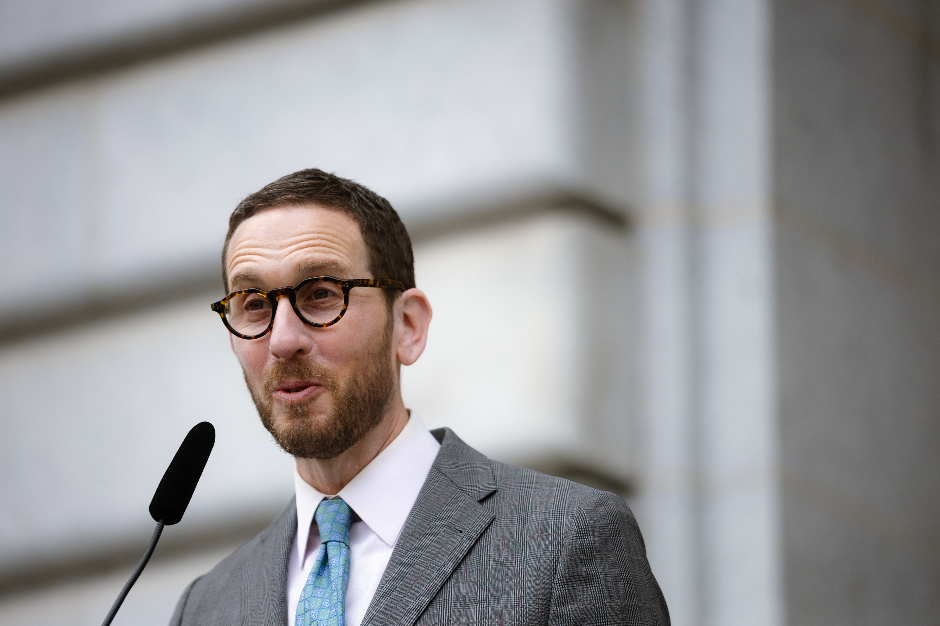 A man in a gray suit and blue tie speaks into a microphone. He has short hair, a beard, and is wearing round glasses. The background is a blurred stone wall.