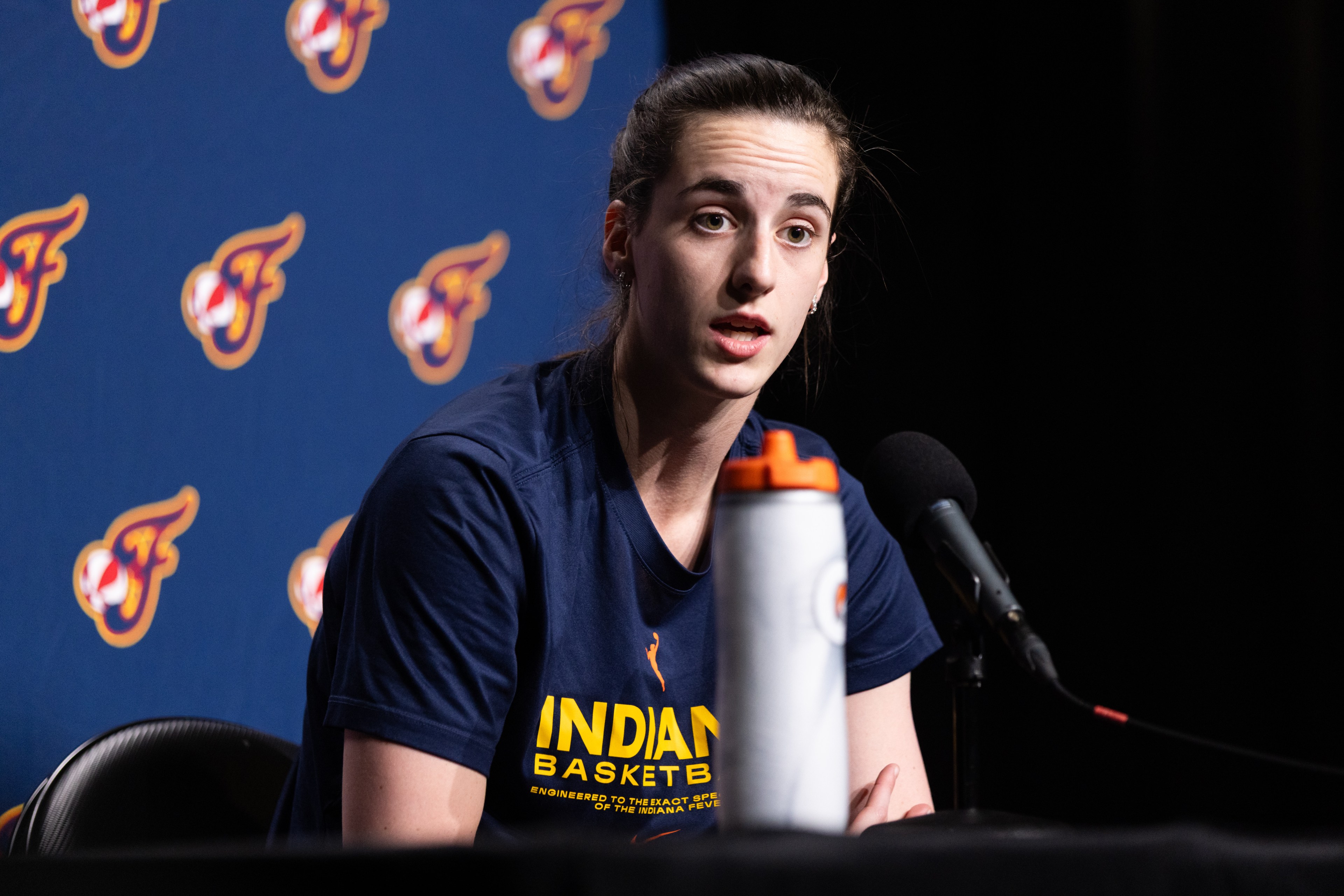 A person is speaking at a press conference, wearing a shirt with &quot;Indiana Basketball&quot; on it. There's a microphone and a water bottle nearby, with a backdrop featuring a logo.