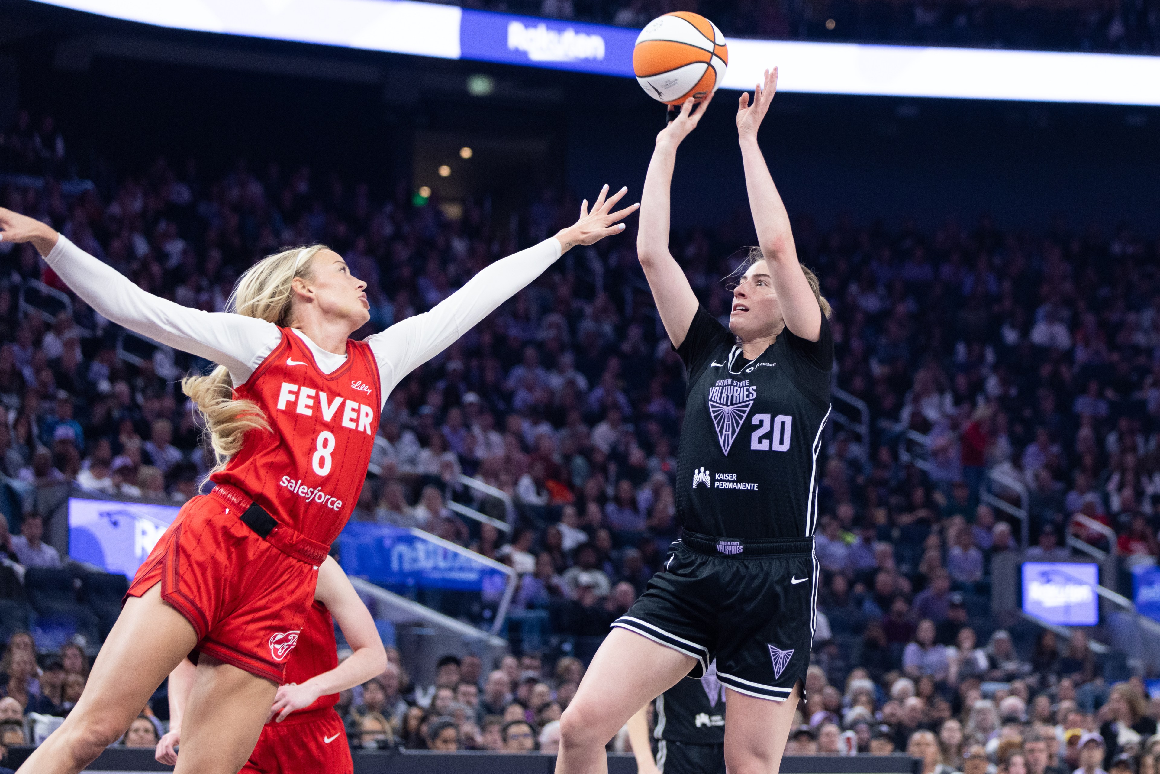 Two basketball players are competing for the ball. One in a red jersey jumps to block, while the other in a black jersey shoots. A crowd watches intently.