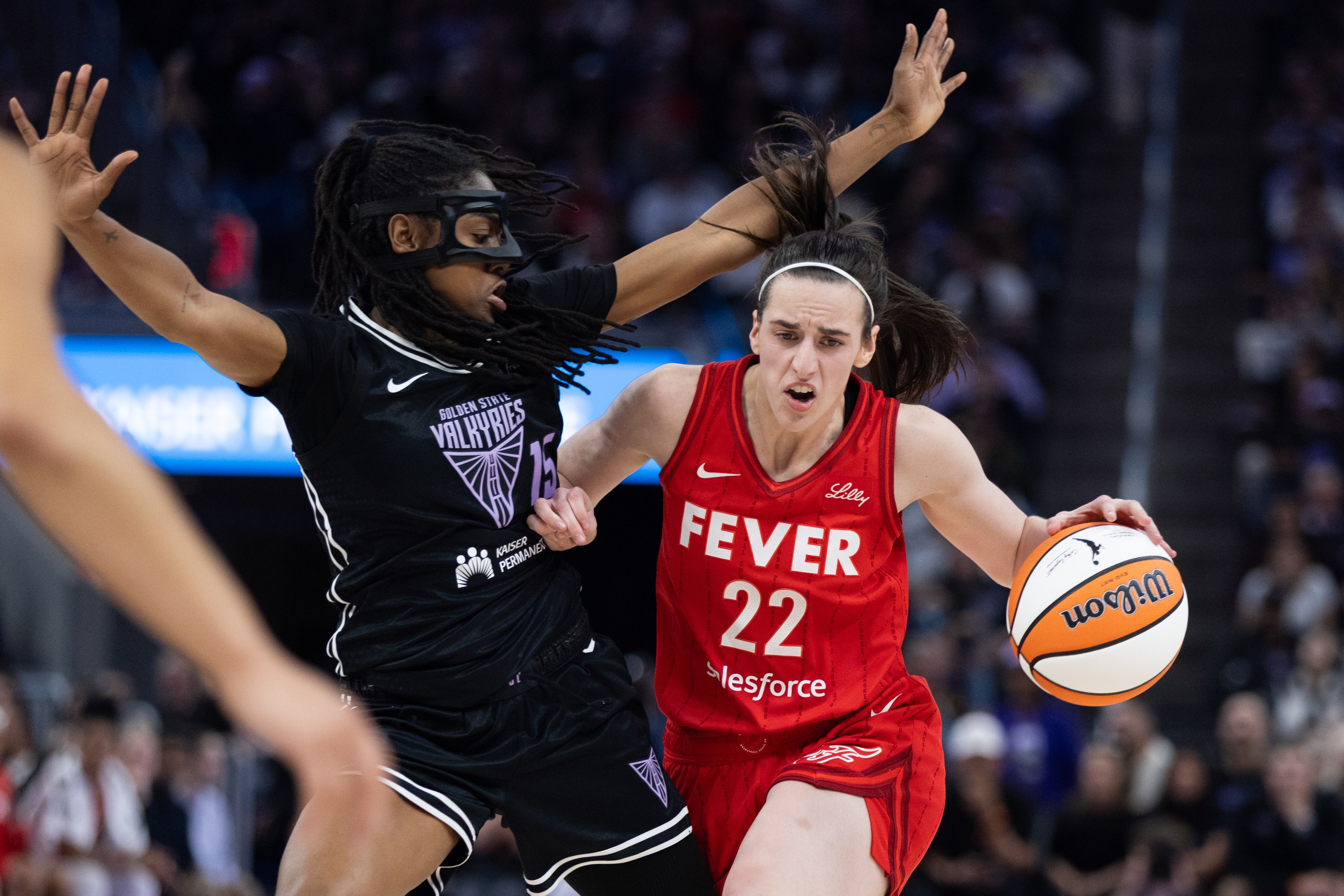 Two basketball players in action, one in a red &quot;FEVER&quot; uniform dribbling, the other in a black &quot;VALKYRIES&quot; uniform defending closely, each showing intense focus.