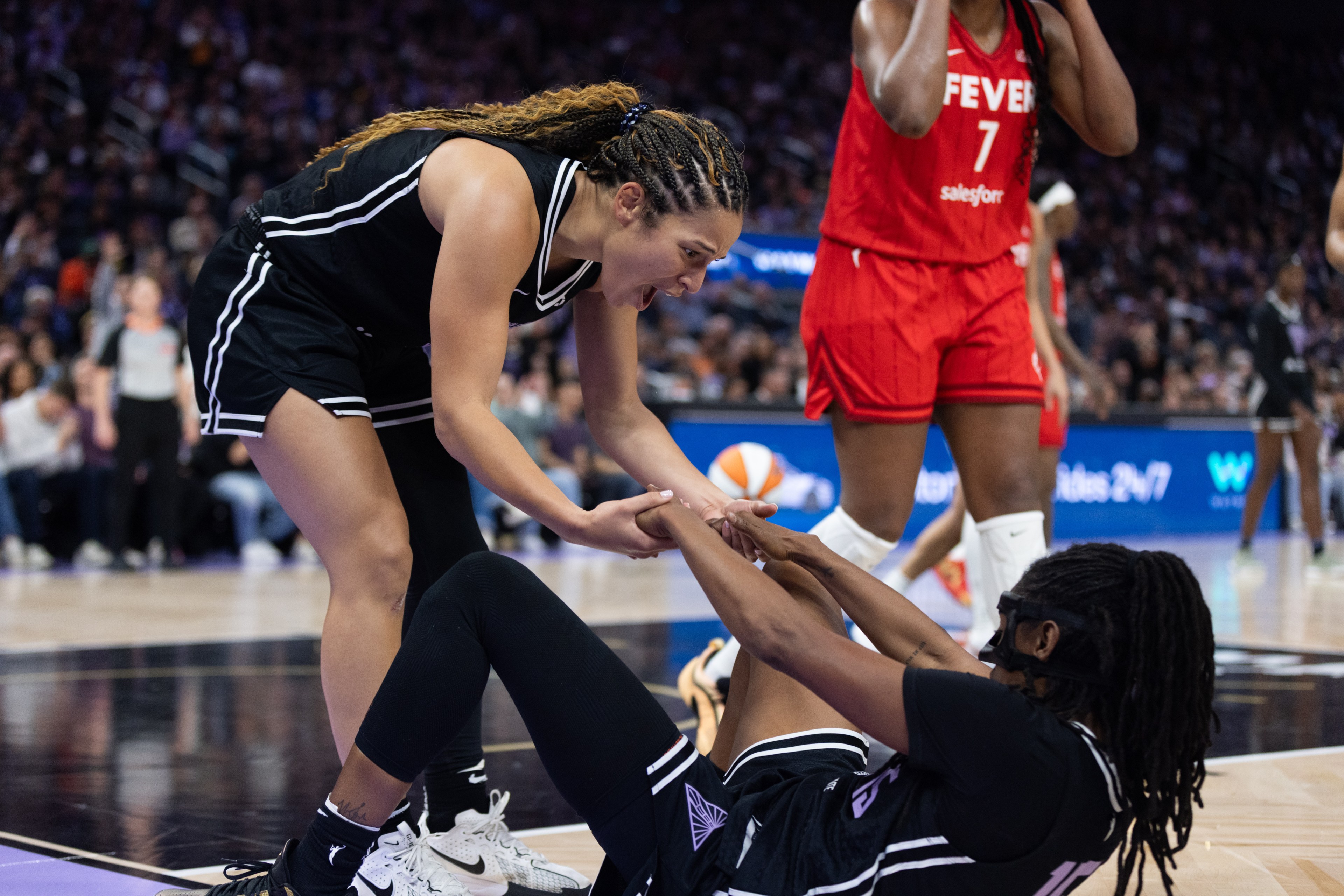 A basketball player in a black uniform helps a teammate up from the floor, with a player in a red uniform nearby. The scene is energetic and supportive.