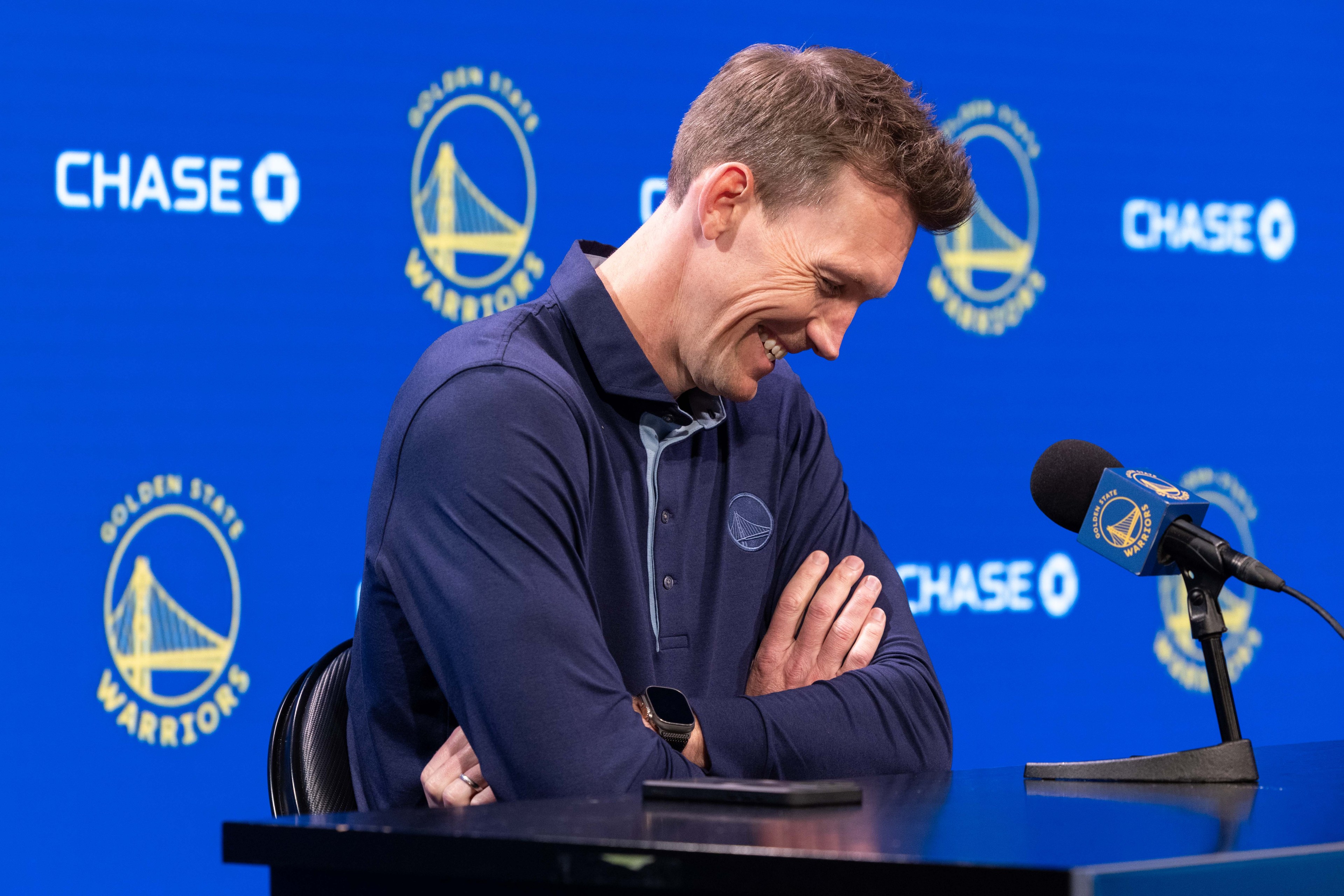 A smiling man stands at a microphone, arms crossed, in front of a blue background with Golden State Warriors logos and the word &quot;CHASE&quot; visible.