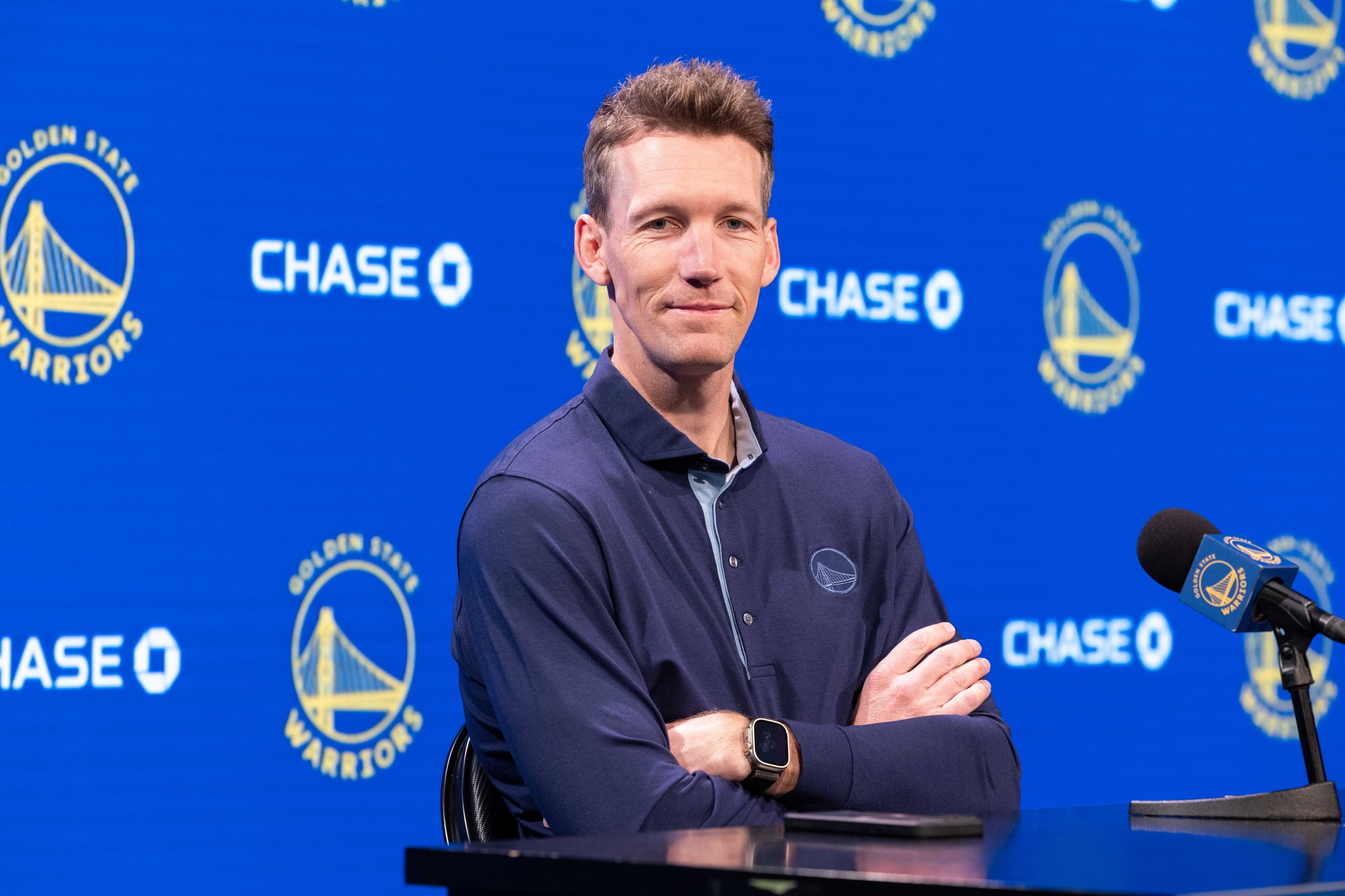 A person in a polo shirt stands with folded arms in front of a blue background adorned with Golden State Warriors logos and "CHASE" text. A microphone is nearby.