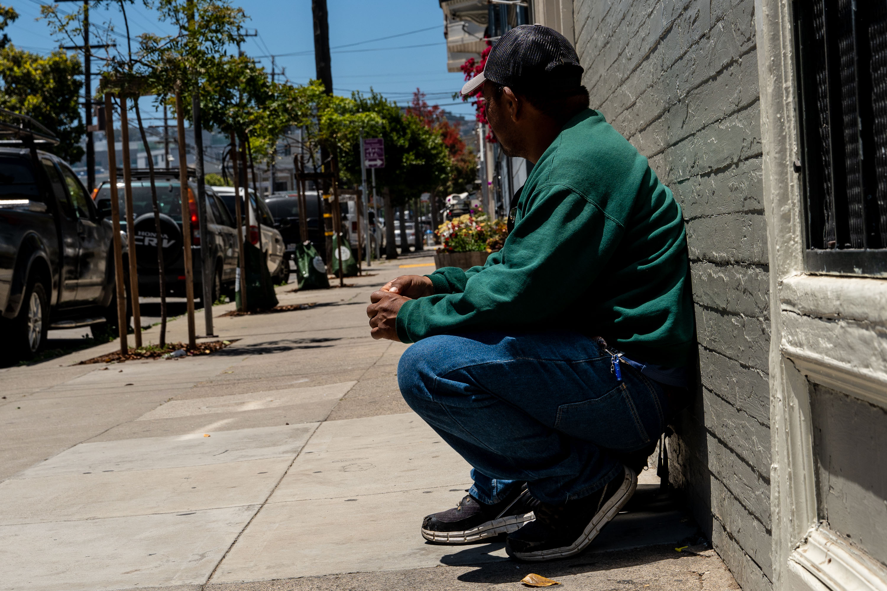 A person in a green sweater and cap is squatting against a brick wall on a sunlit sidewalk. Trees and parked cars line the street ahead.