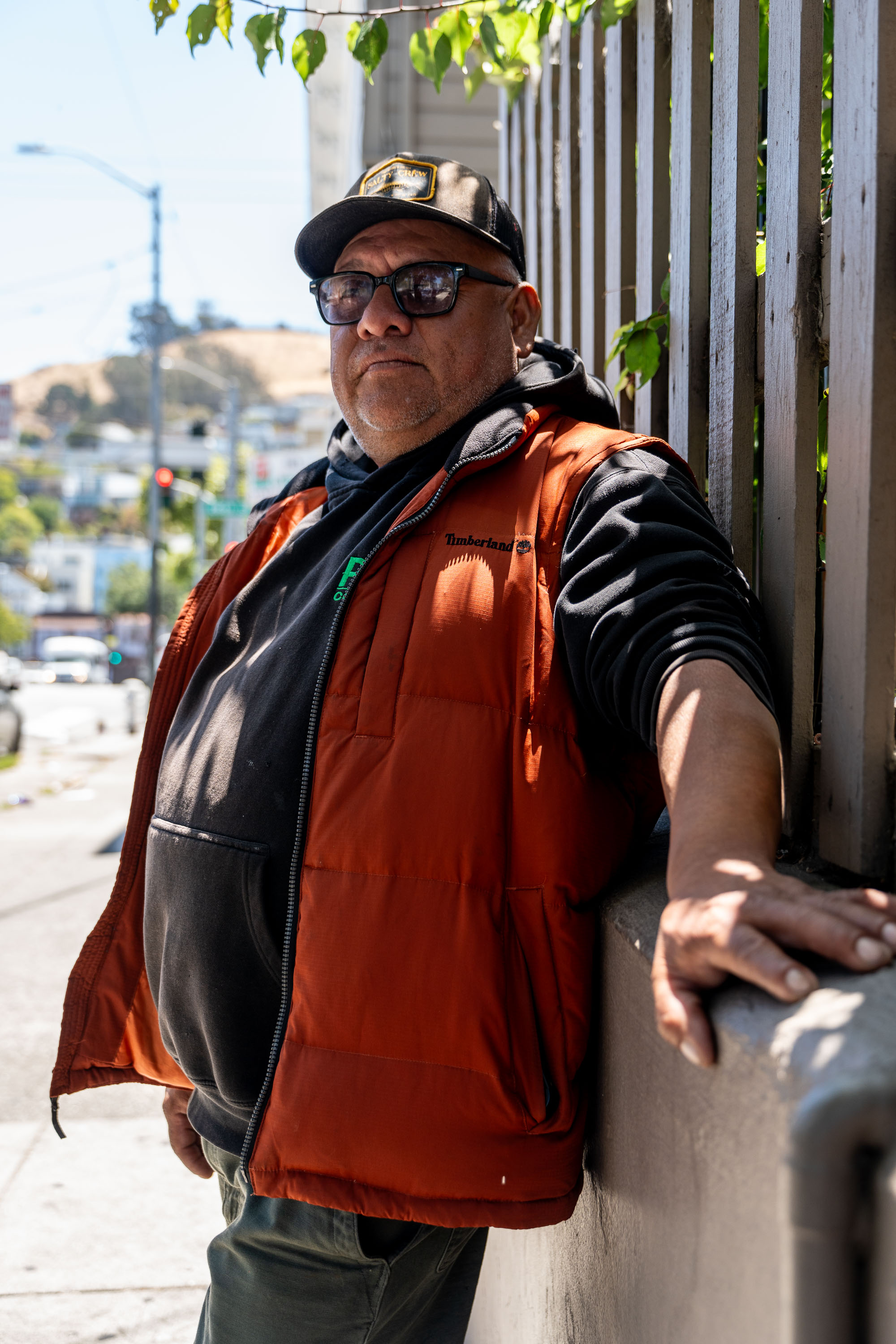 A man leans on a railing outdoors, wearing sunglasses, a black cap, an orange vest, and a black hoodie. The sun casts shadows, and trees are in the background.