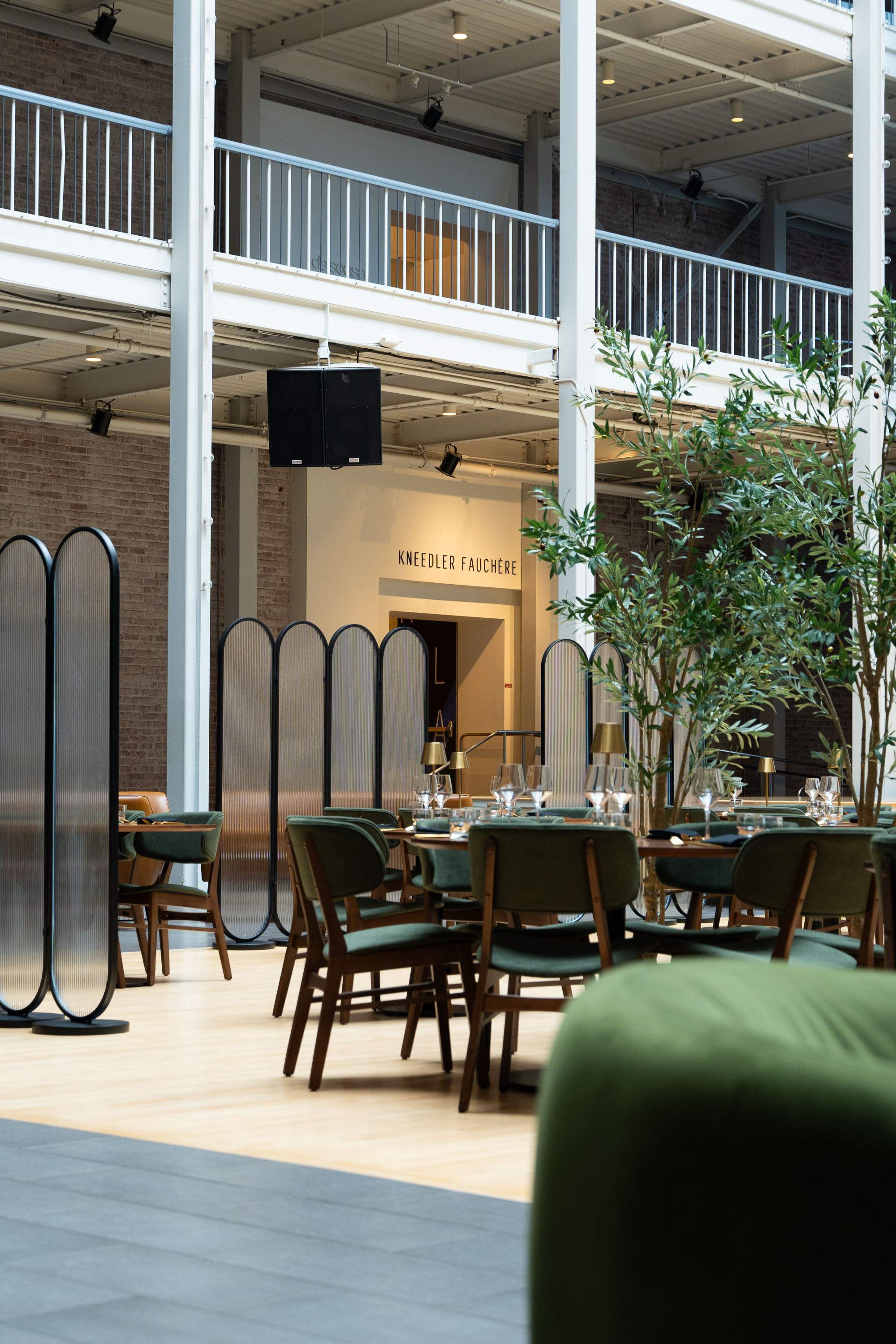 The image shows a modern dining space with green chairs, neatly set tables, indoor plants, and a bright atmosphere. A sign above reads &quot;KNEEDLER FAUCHÈRE.&quot;