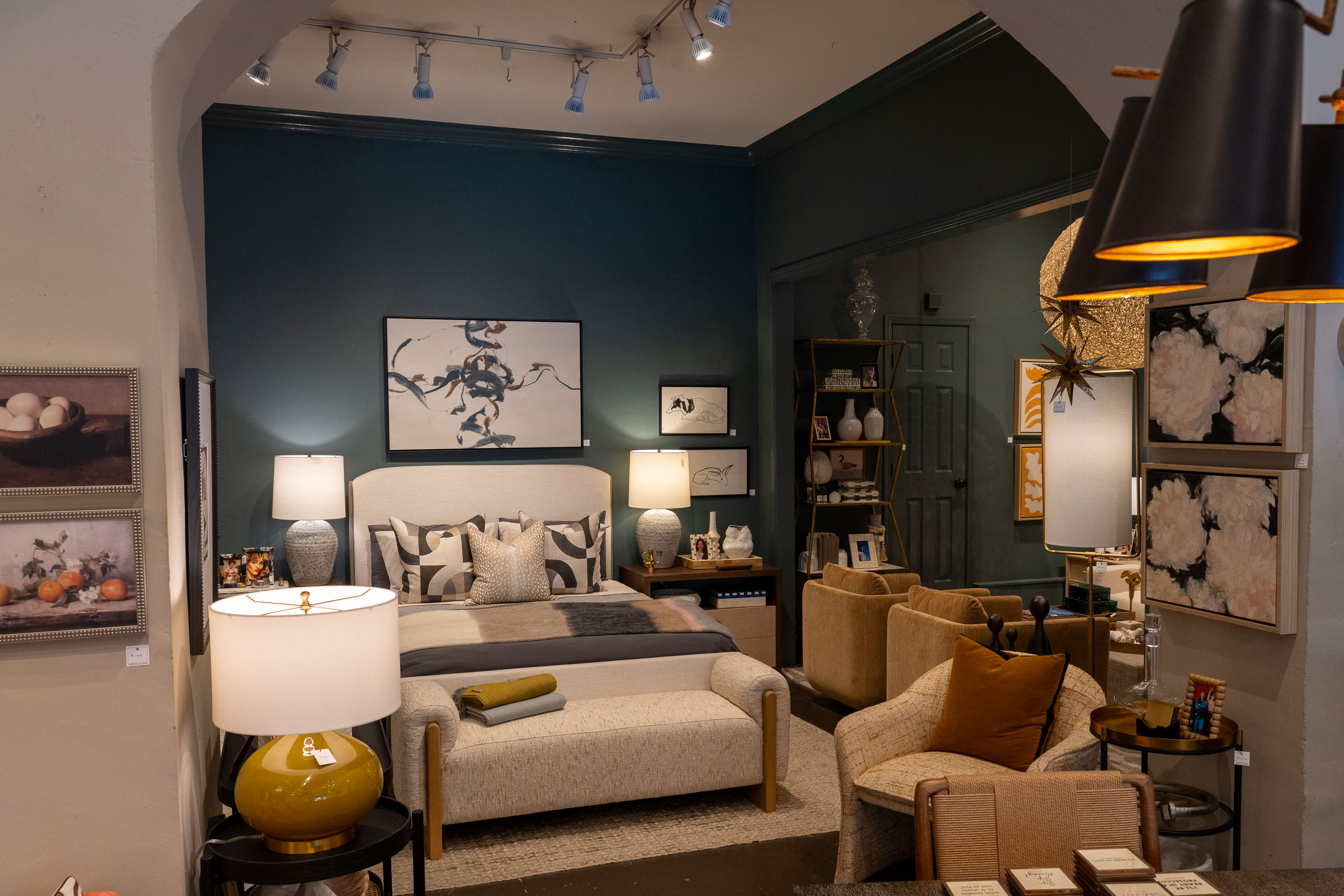 A cozy room with a beige bed, decorative pillows, and various art pieces on blue and beige walls. Soft lighting and warm-toned furniture complete the space.