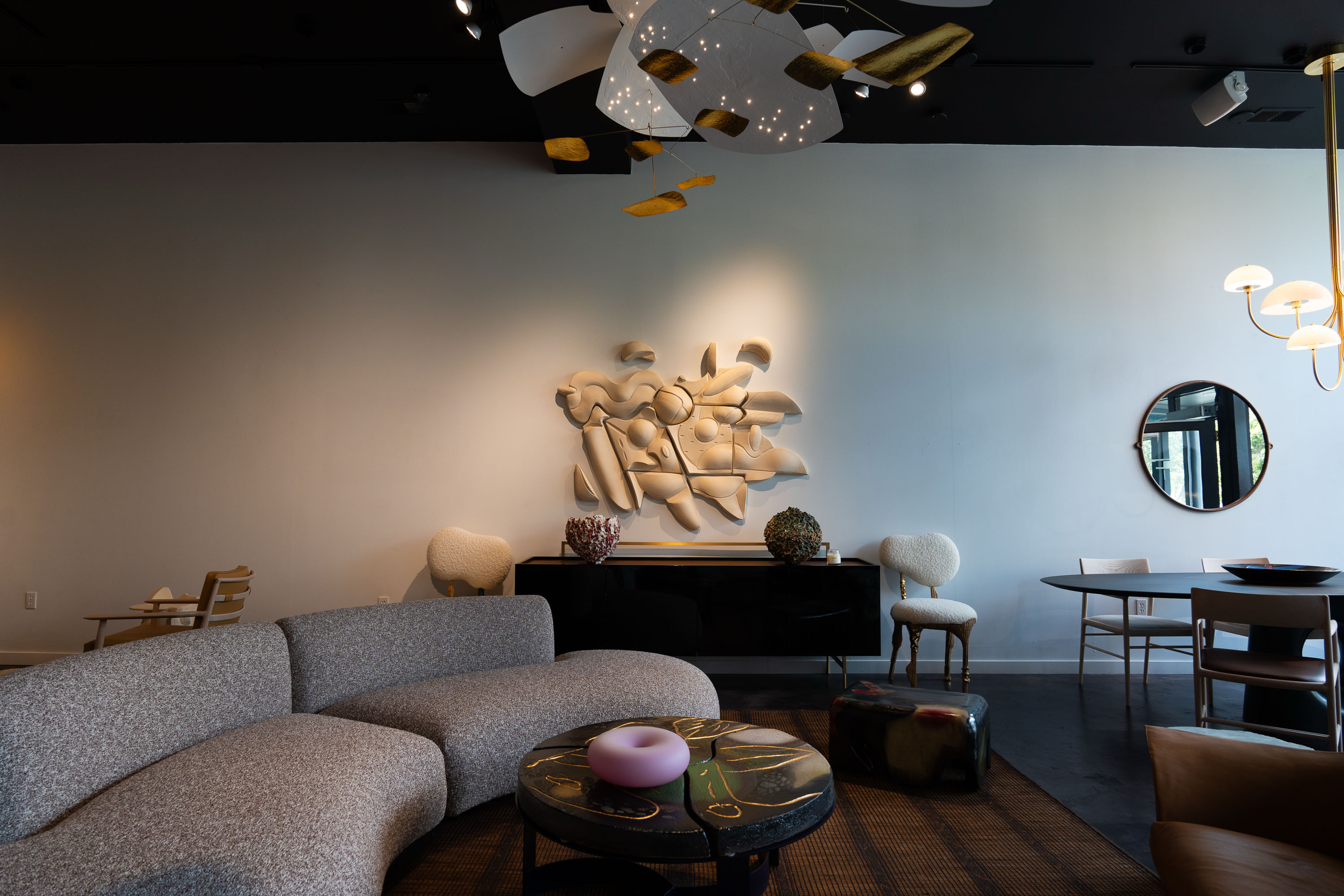 The image shows a modern, stylish living room with a curved gray sofa, abstract wall art, a circular coffee table, chairs, a round mirror, and a unique ceiling light.