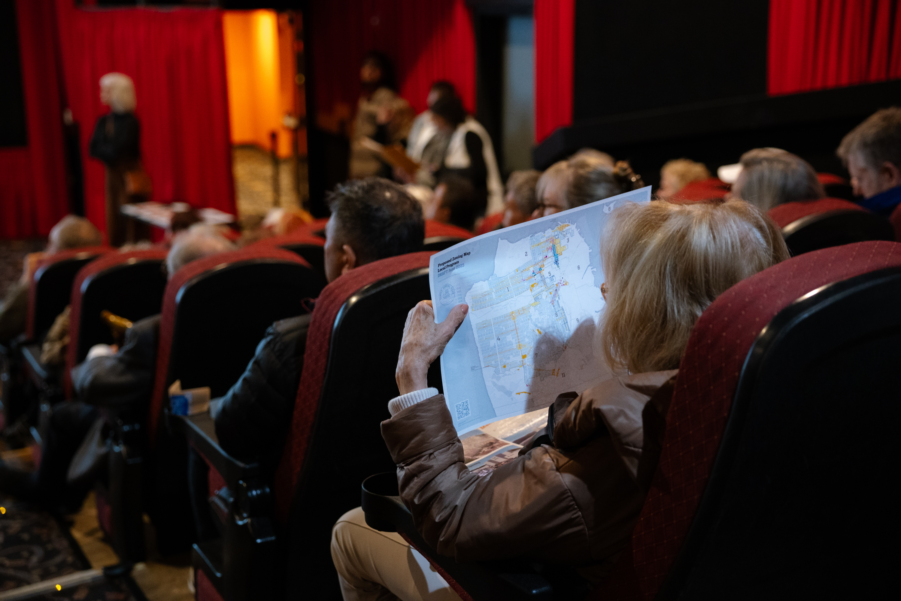 People are sitting in a theater with red seats, attentive to a speaker. One person is holding a map, while others face forward, focused on the stage.