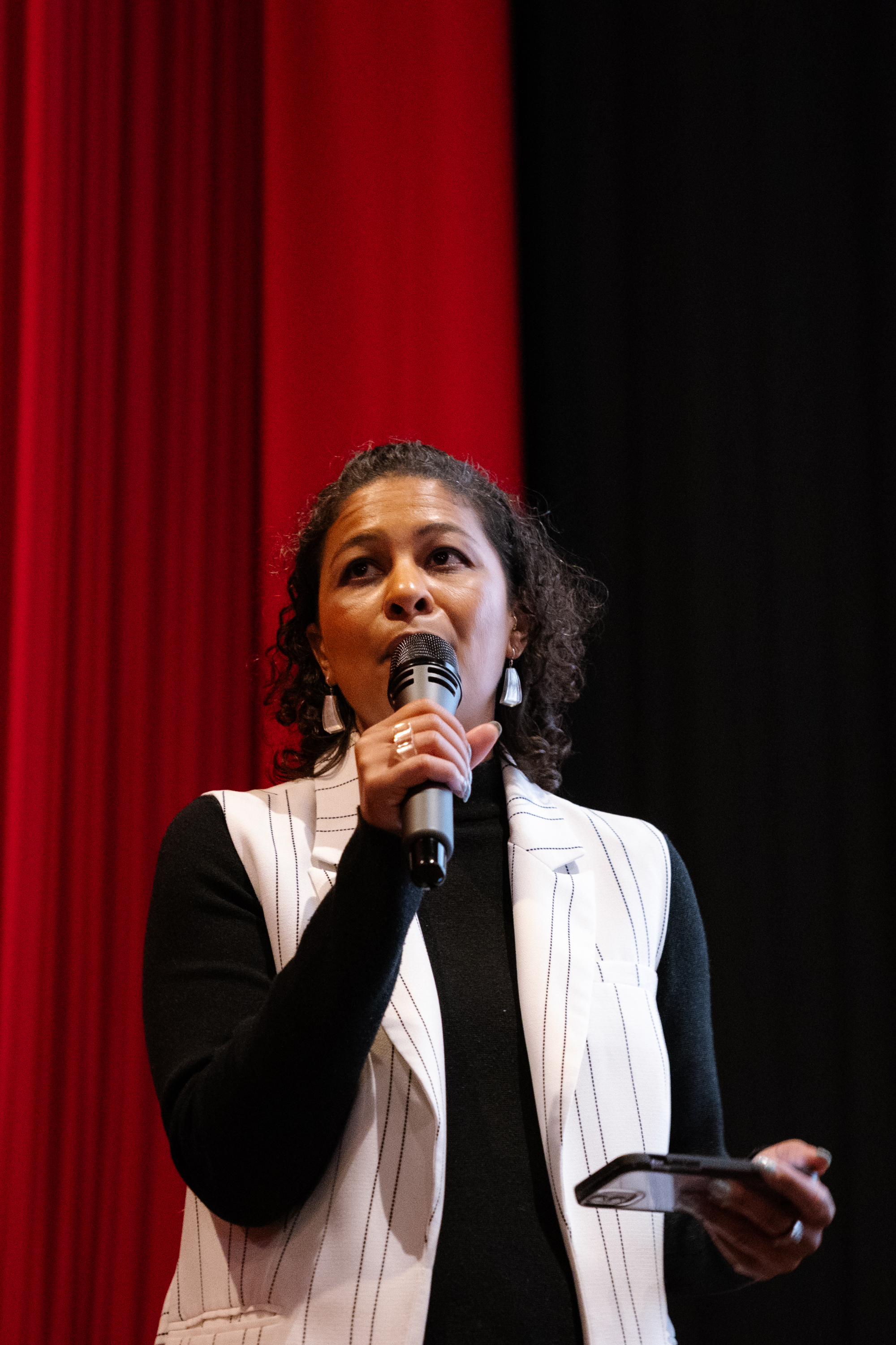 A person stands holding a microphone, speaking. They are wearing a light-colored vest over a dark top, with red and black curtains in the background.
