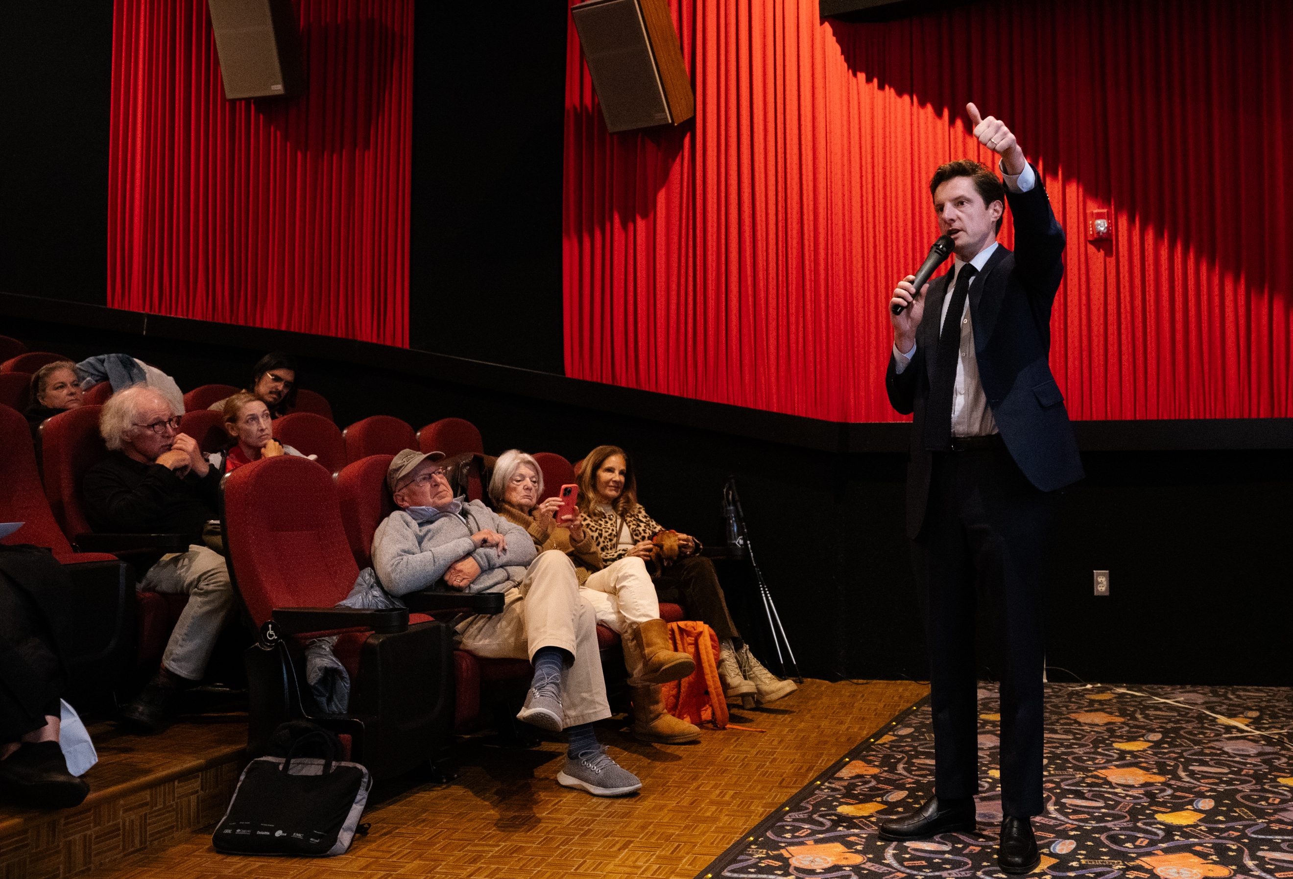 A man in a suit speaks into a microphone, gesturing with a thumbs-up, in a theater with red seats and walls. An audience watches attentively.