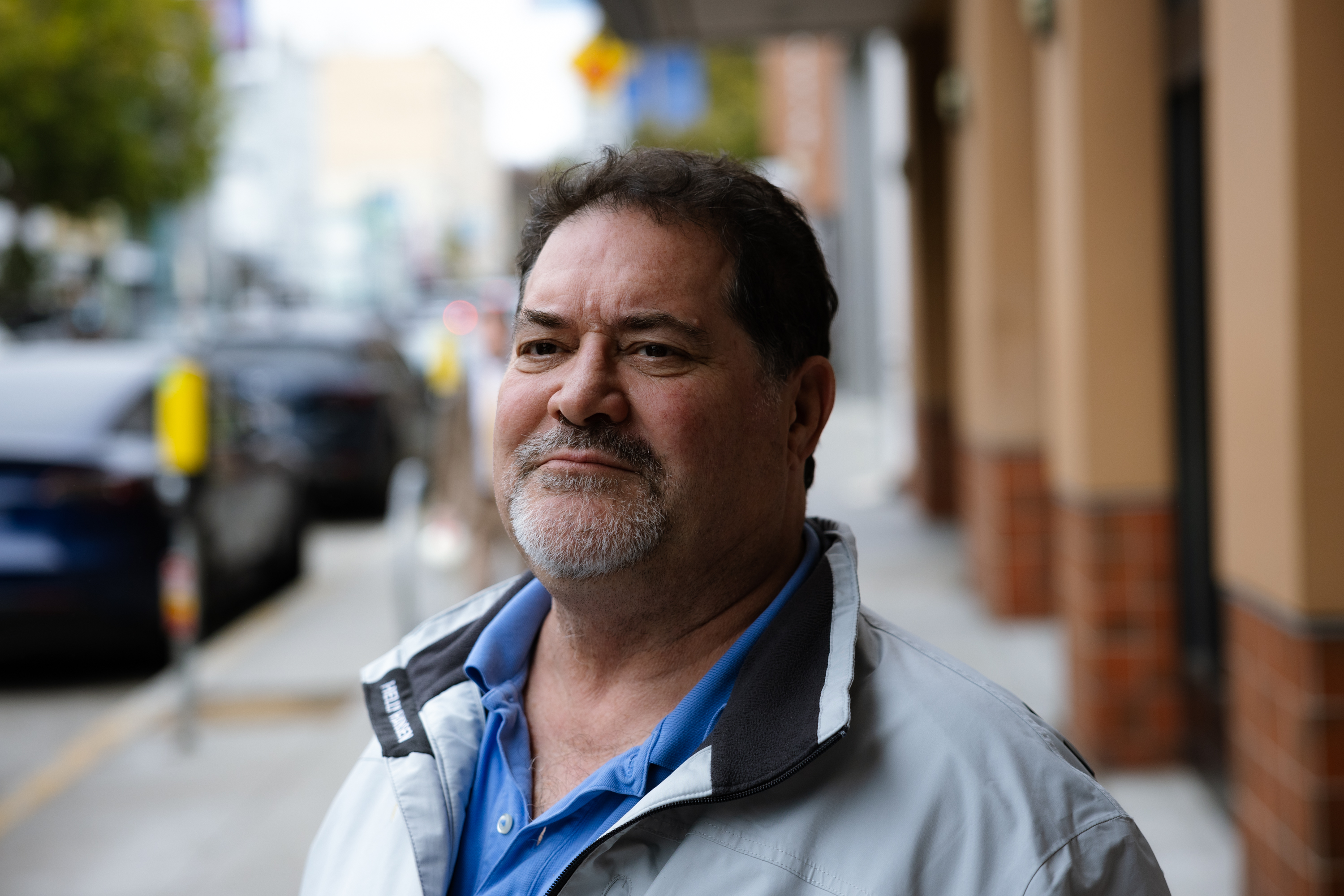 A middle-aged man with a beard smiles slightly while standing on a city sidewalk. He wears a light jacket over a blue shirt. The background is blurred.