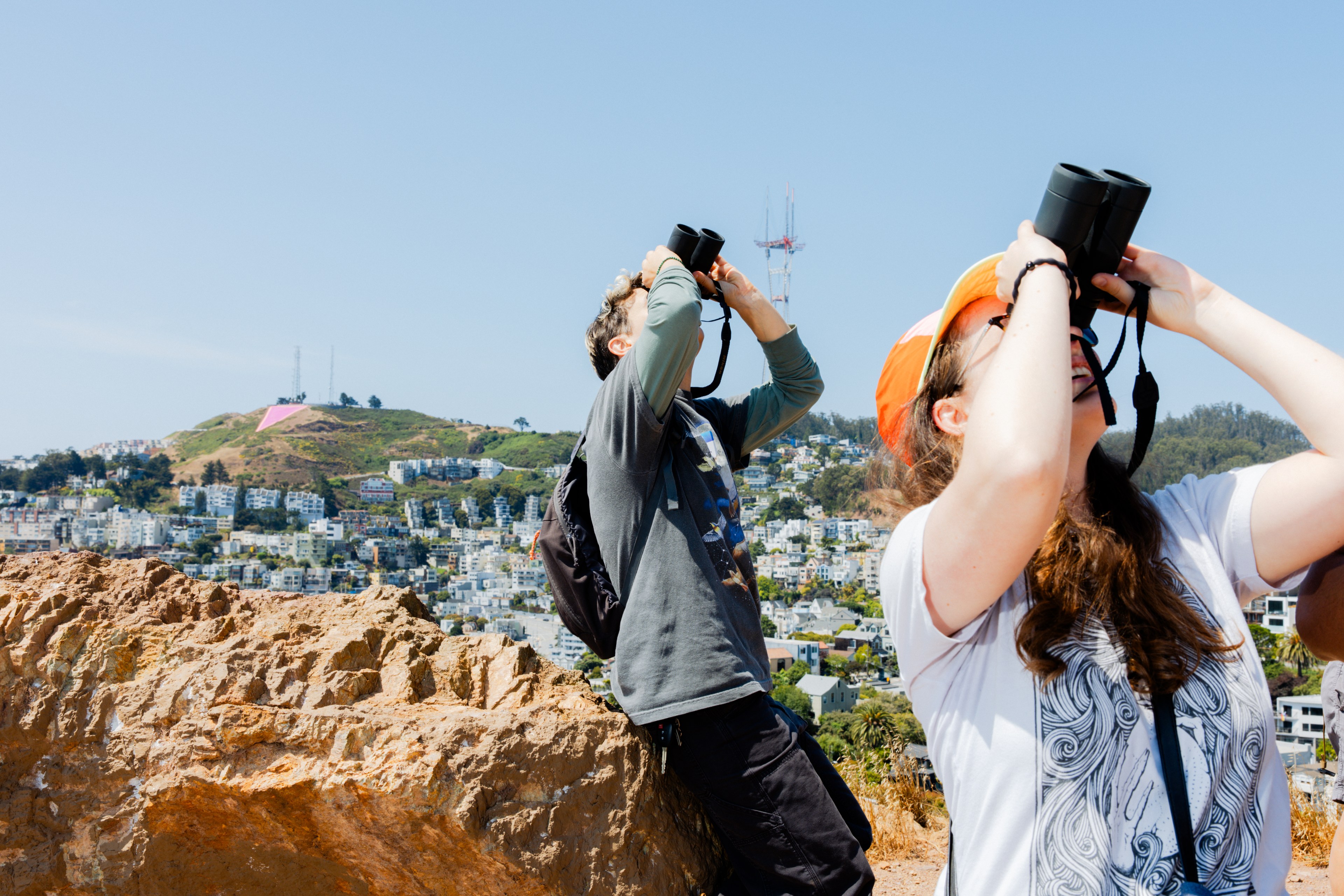 Two people stand on a rocky hilltop using binoculars, with a cityscape and greenery in the background. One wears an orange cap and the other a backpack.