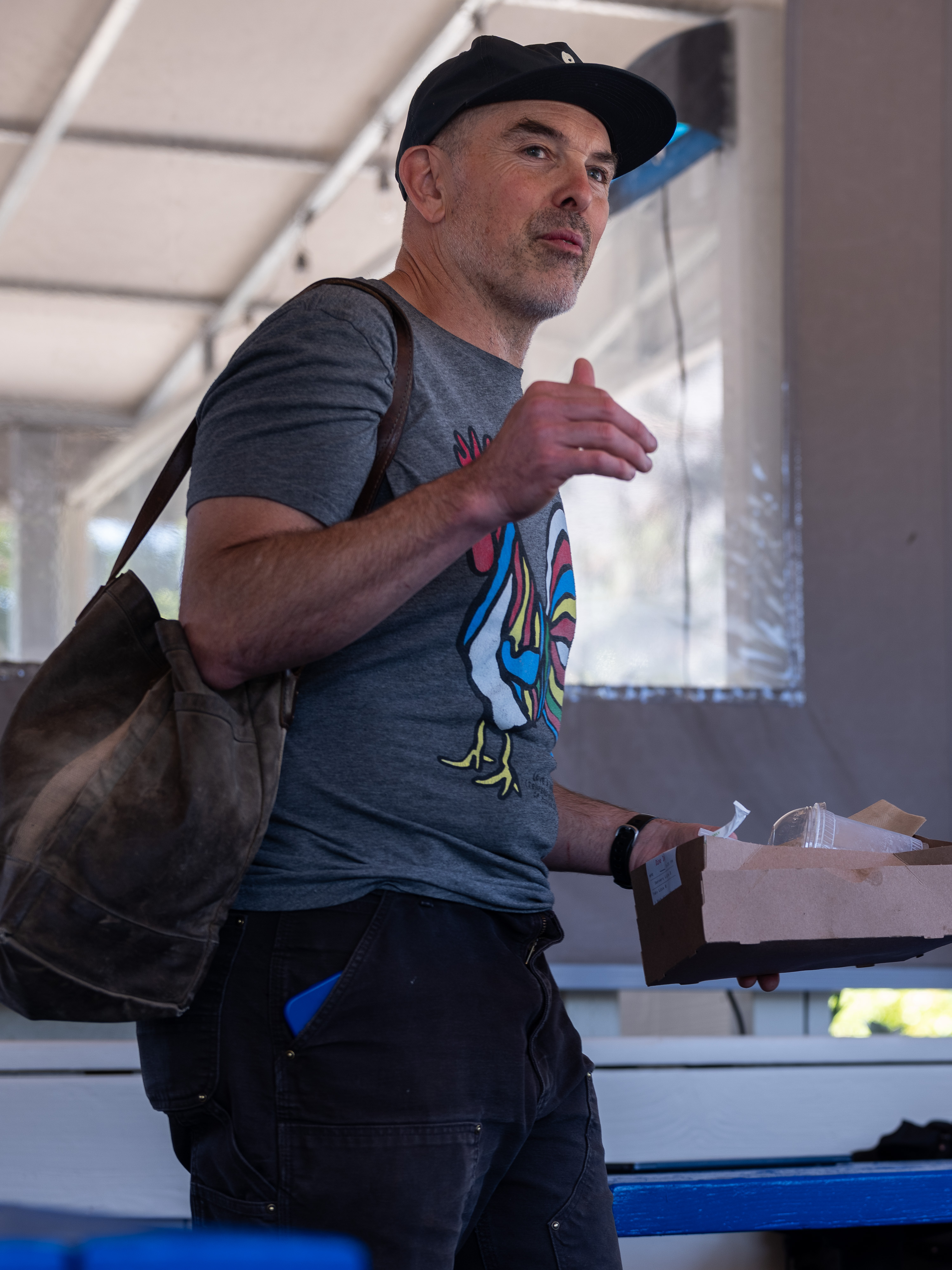 A person in a cap and gray rooster T-shirt holds a brown paper tray. They have a bag over one shoulder and stand indoors with blurred background.