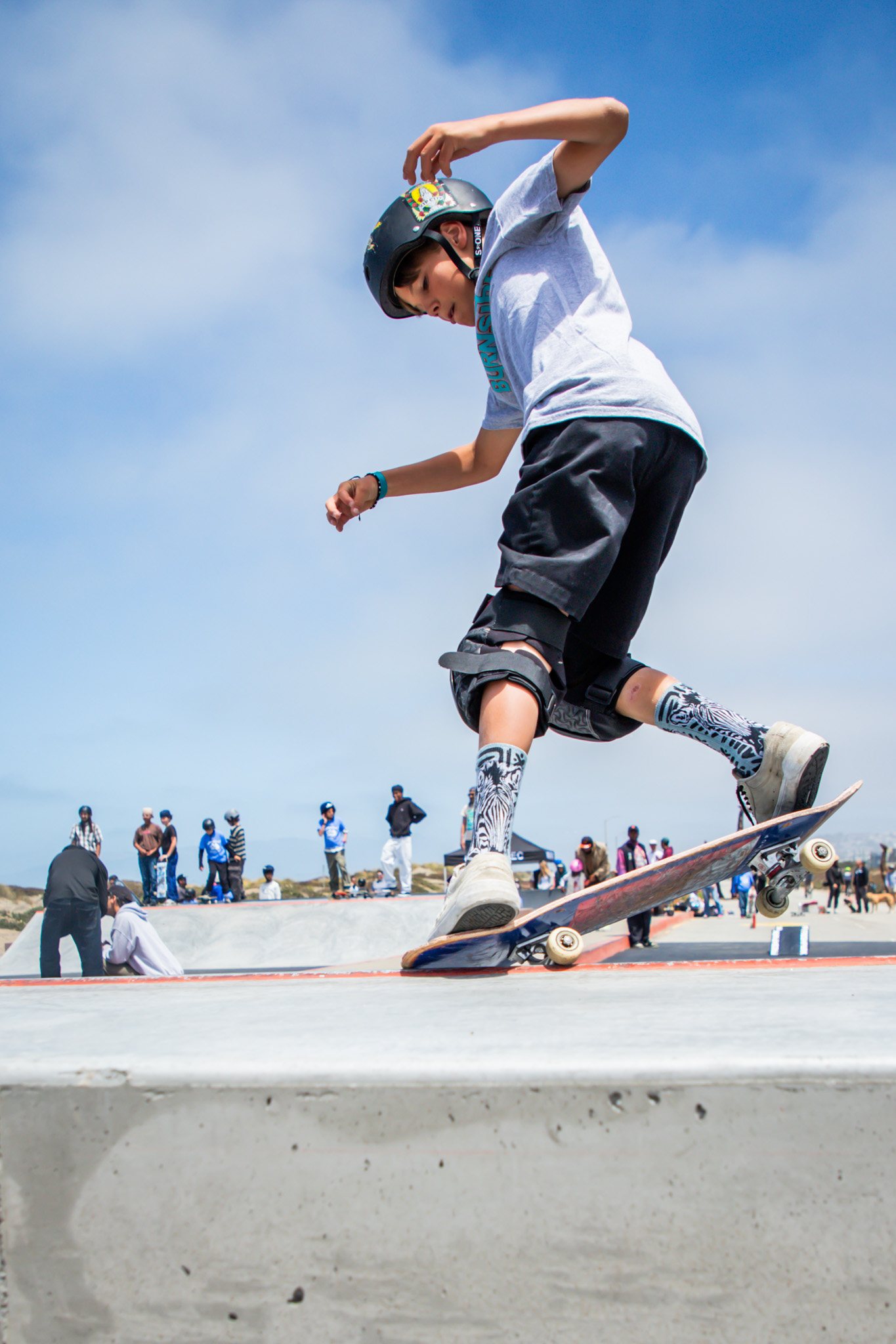 A young skateboarder in a helmet performs a trick on a ramp. He wears a gray shirt, black shorts, and patterned socks. People watch in the background.
