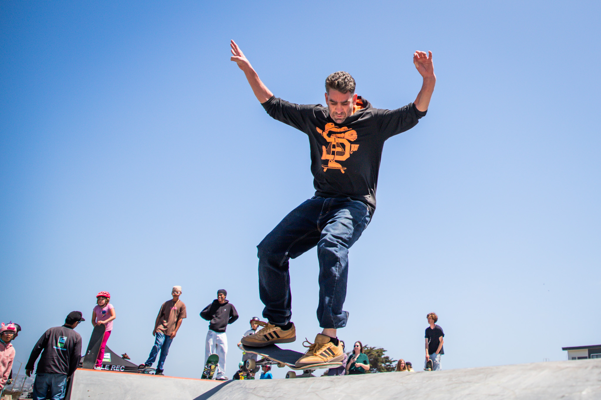 A man in a black hoodie and jeans is skateboarding on a ramp, mid-trick, with his arms out. Behind him, several people, including kids with helmets, watch.