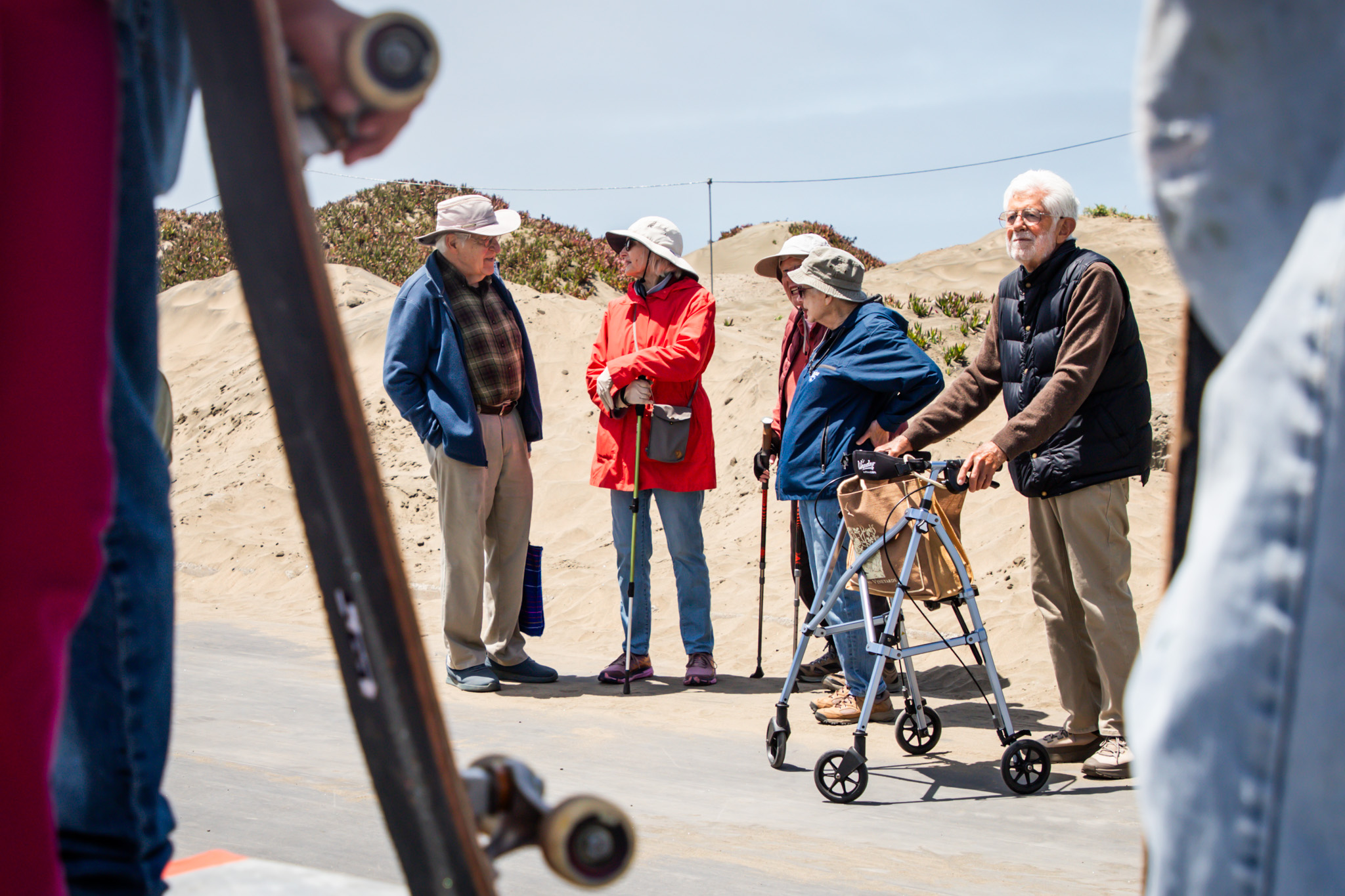 Five elderly people stand outdoors, chatting. Two use walking aids and all wear jackets and hats. They are surrounded by sand dunes, with skateboards nearby.