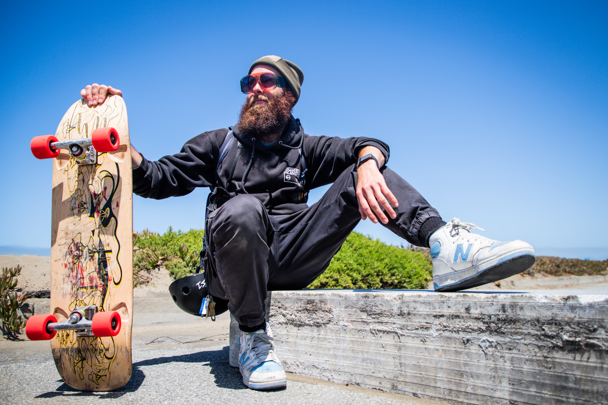 The image shows a bearded person in sunglasses and a beanie sitting on a ledge, holding a skateboard with red wheels. The background is a bright, sunny day.