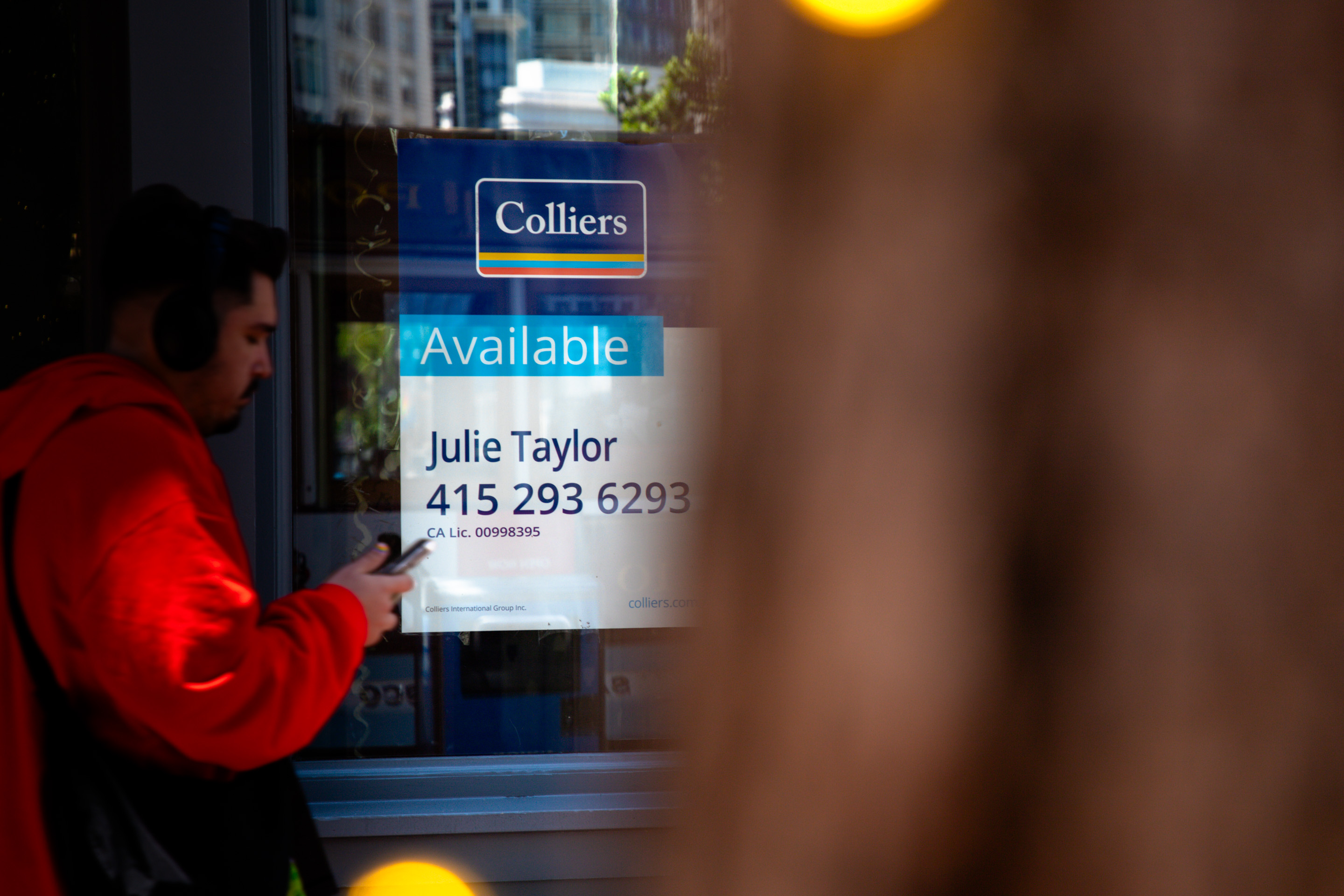 A person in a red hoodie uses a phone near a window displaying a Colliers real estate sign with contact details for an available property.
