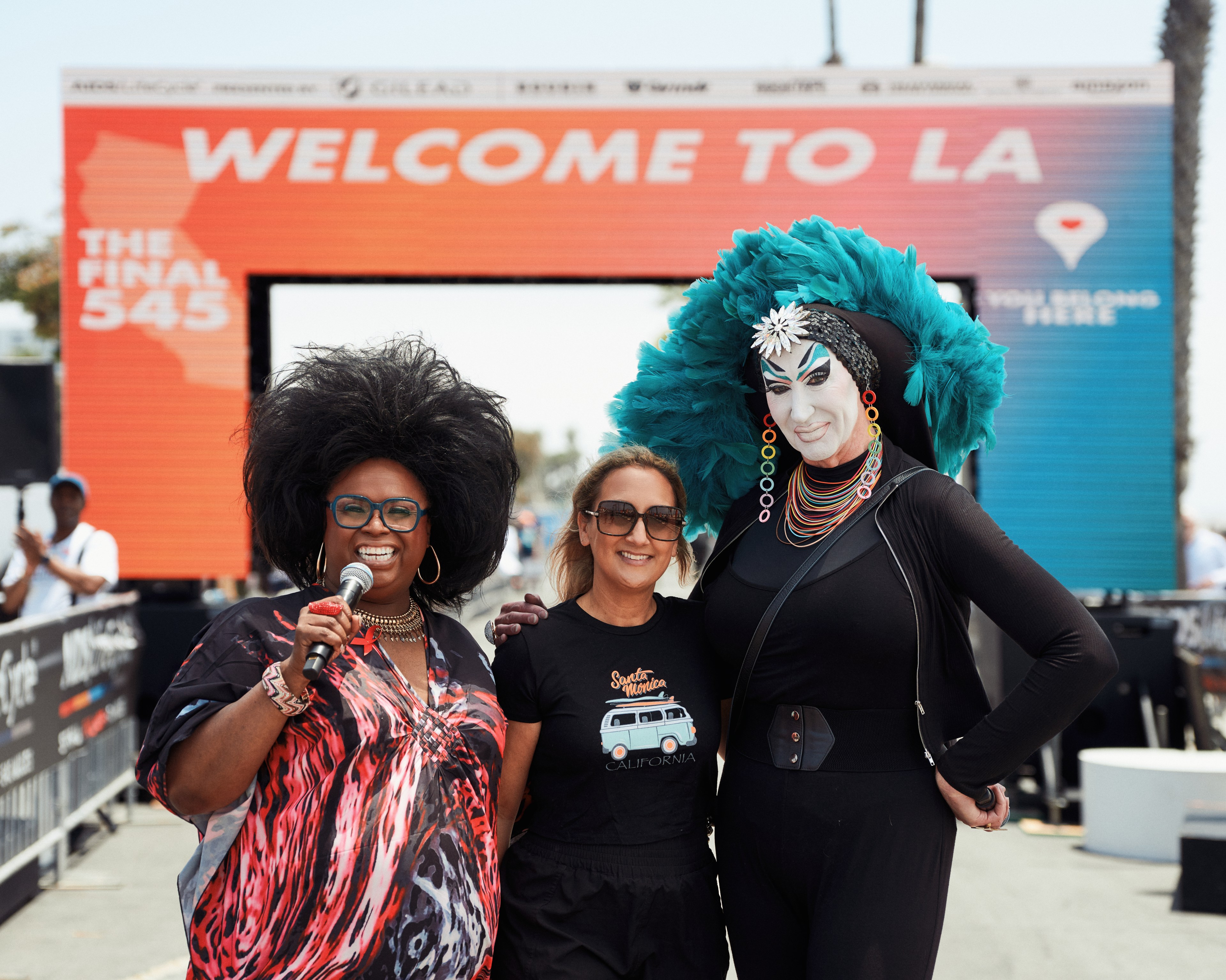 Three people pose in front of a &quot;Welcome to LA&quot; sign. One holds a microphone, another wears a hat with blue feathers, and the third has sunglasses, all smiling.