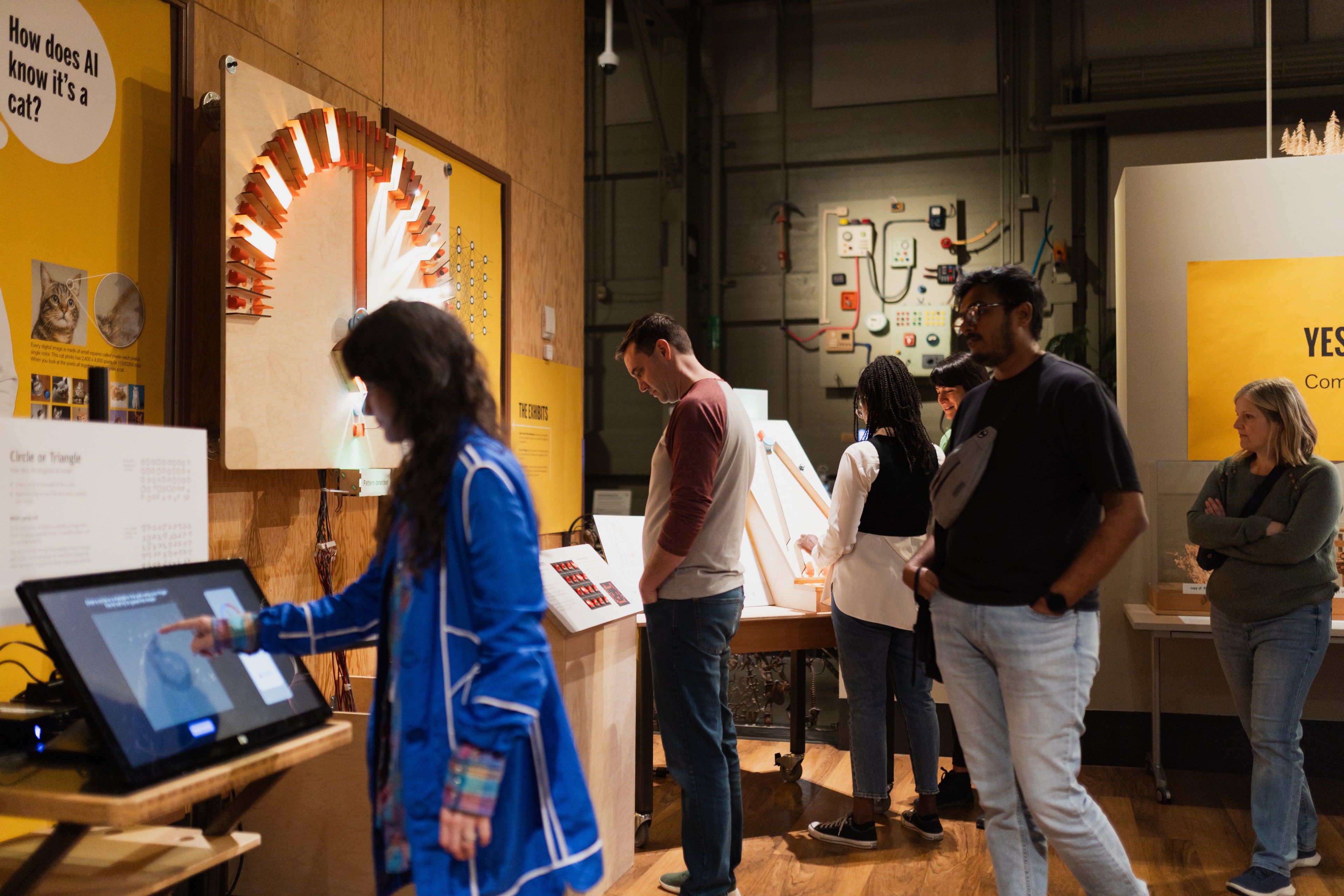 The image shows several people in an interactive exhibit about AI. They engage with displays, a touchscreen, and panels, exploring various concepts.