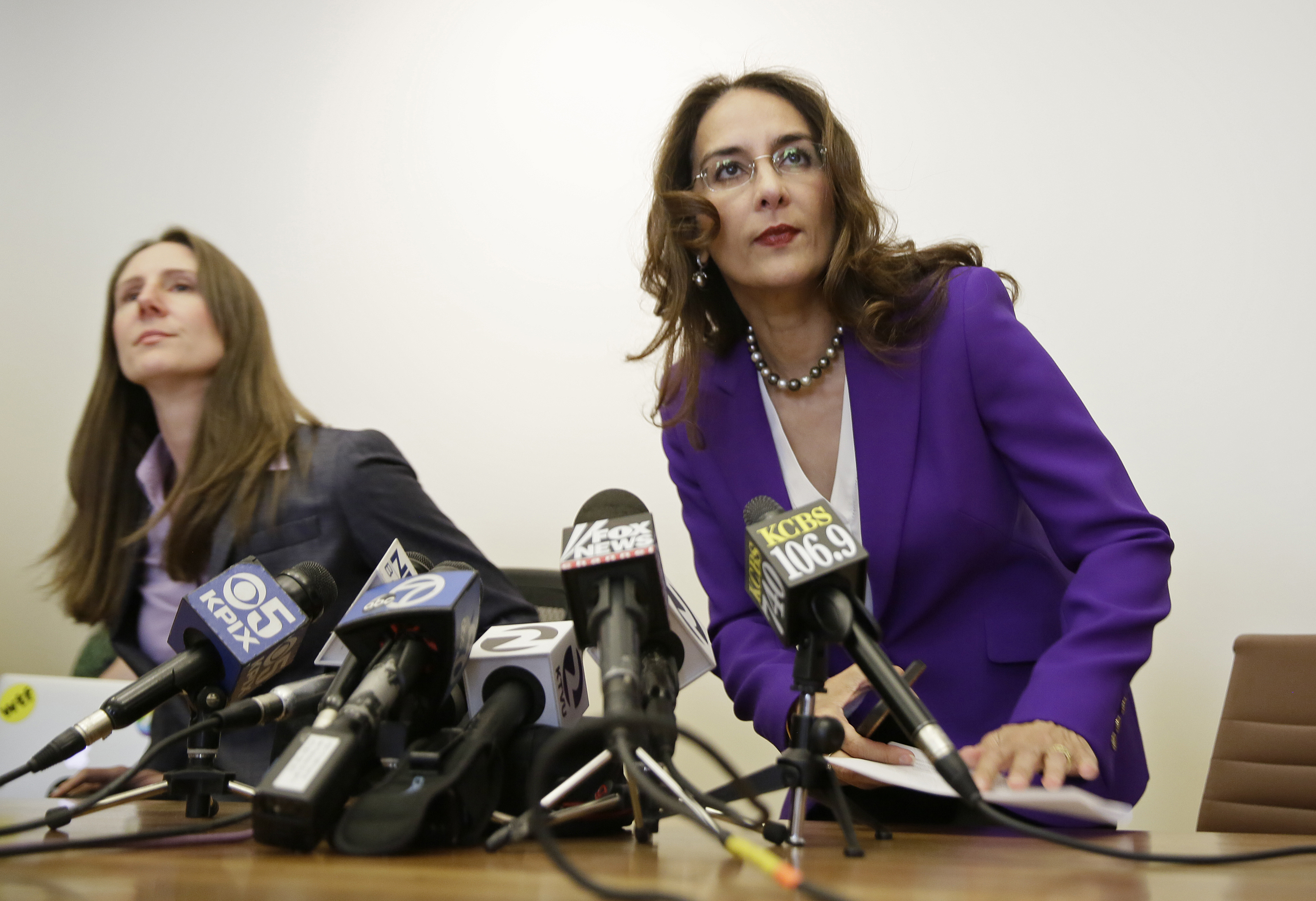Two women are seated at a table with multiple microphones in front of them, possibly preparing for a press conference. One woman wears a purple suit.