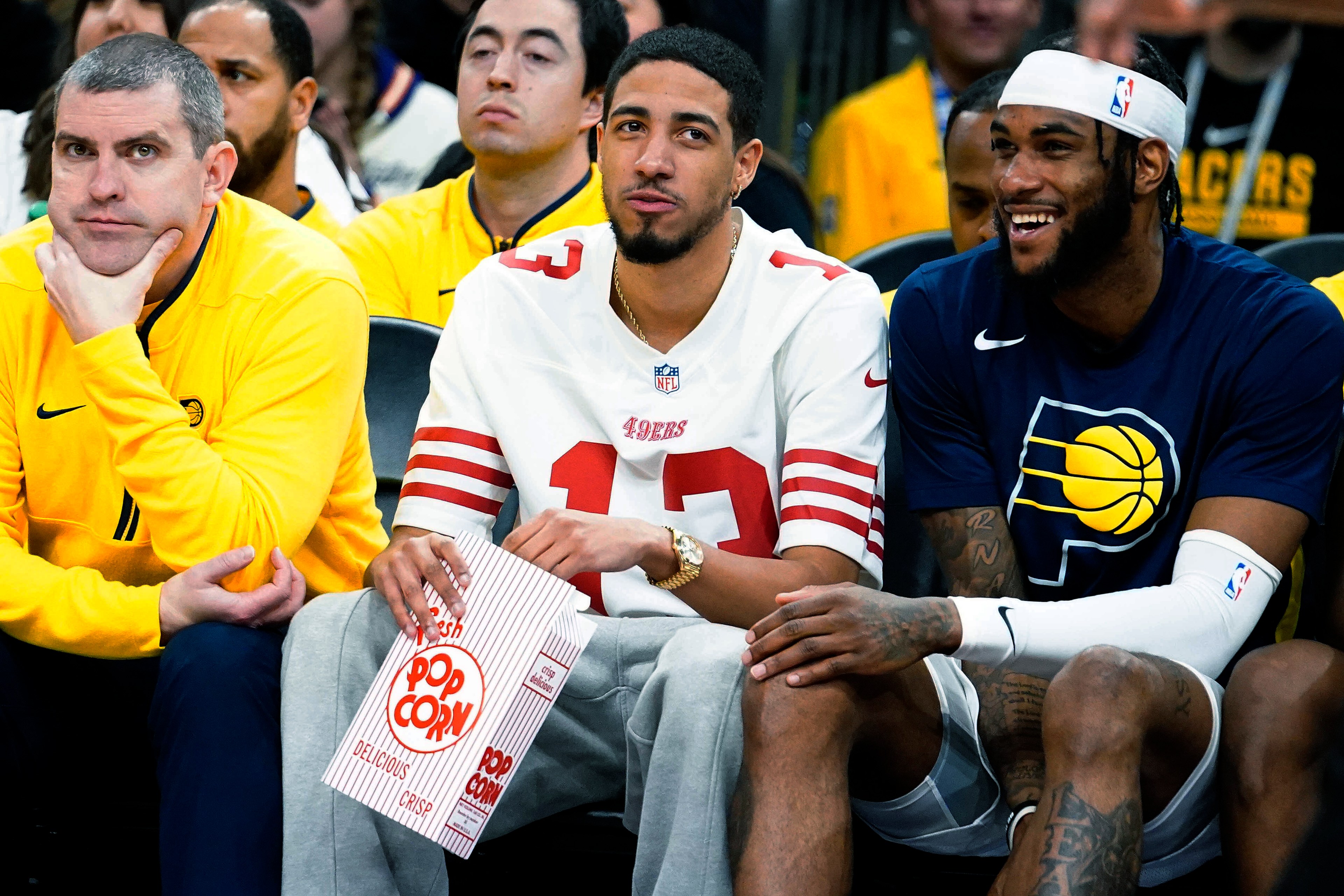 Two men are seated at a sports event, one in a 49ers jersey holding popcorn, the other in a Pacers shirt, smiling. People in yellow shirts surround them.