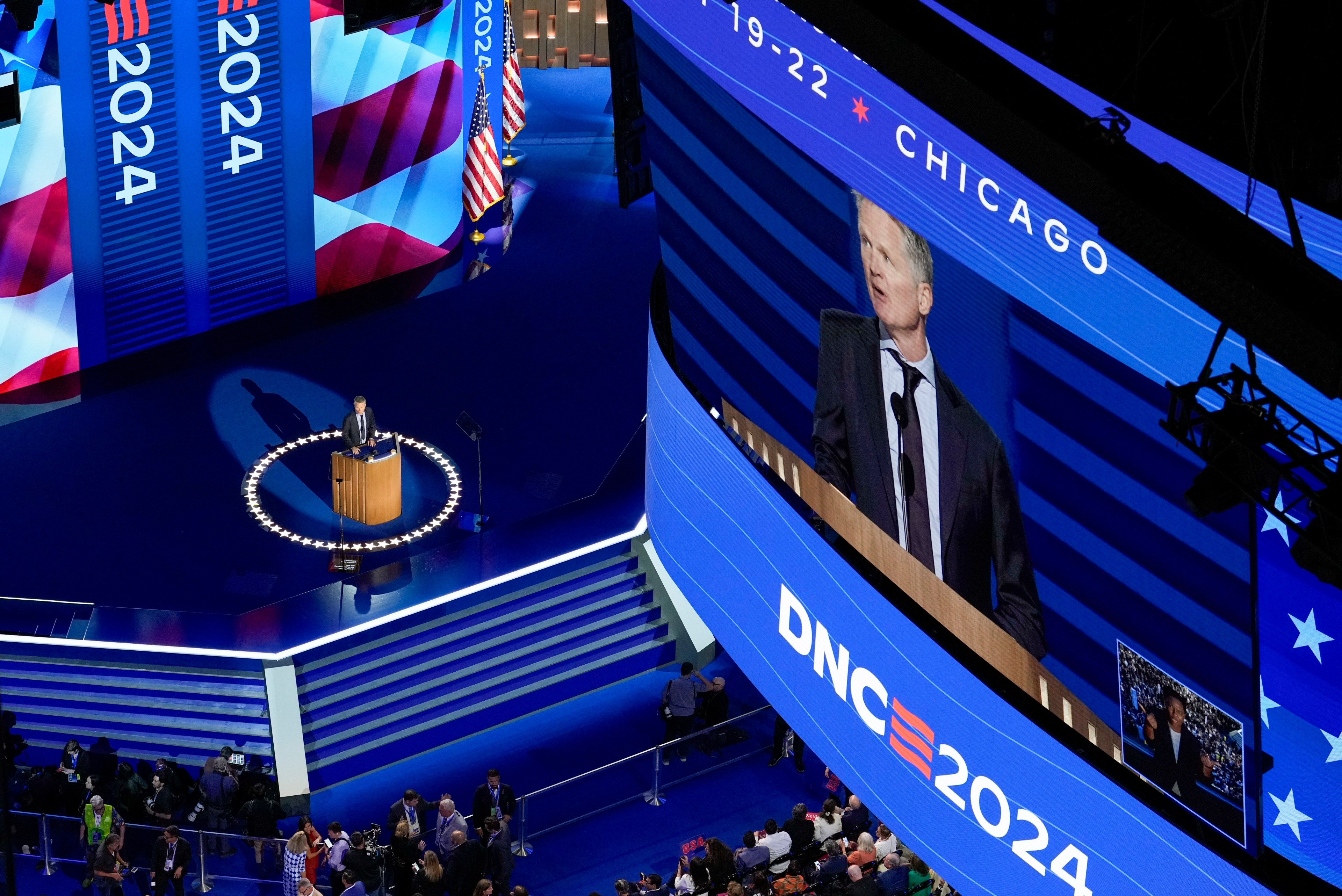 A speaker stands at a podium on a blue-lit stage, surrounded by stars. &quot;DNC 2024&quot; is displayed on screens, with an audience below and American flags visible.