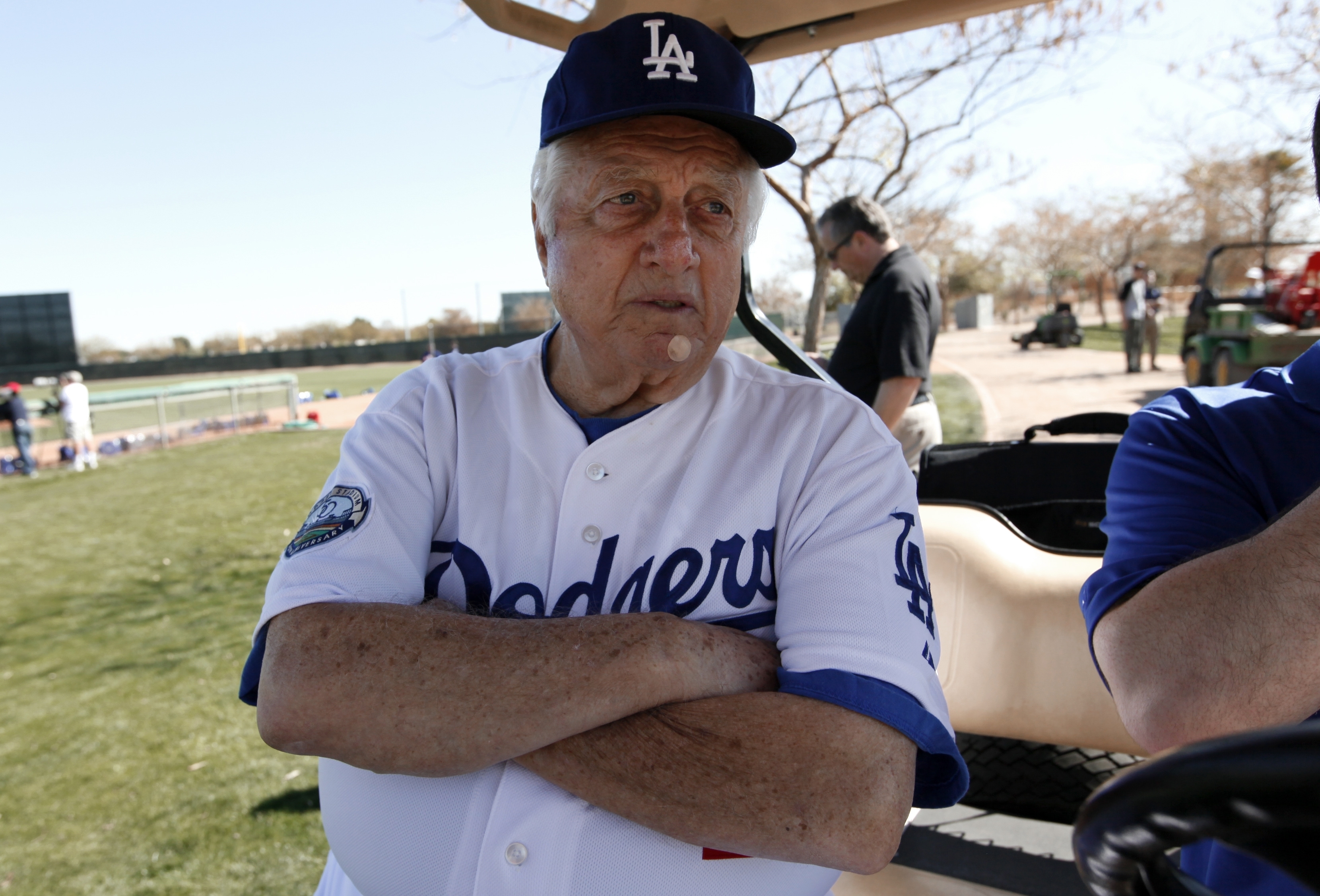 An older man in a Dodgers baseball uniform stands with crossed arms near a sports field, wearing a cap. There are people and trees in the background.
