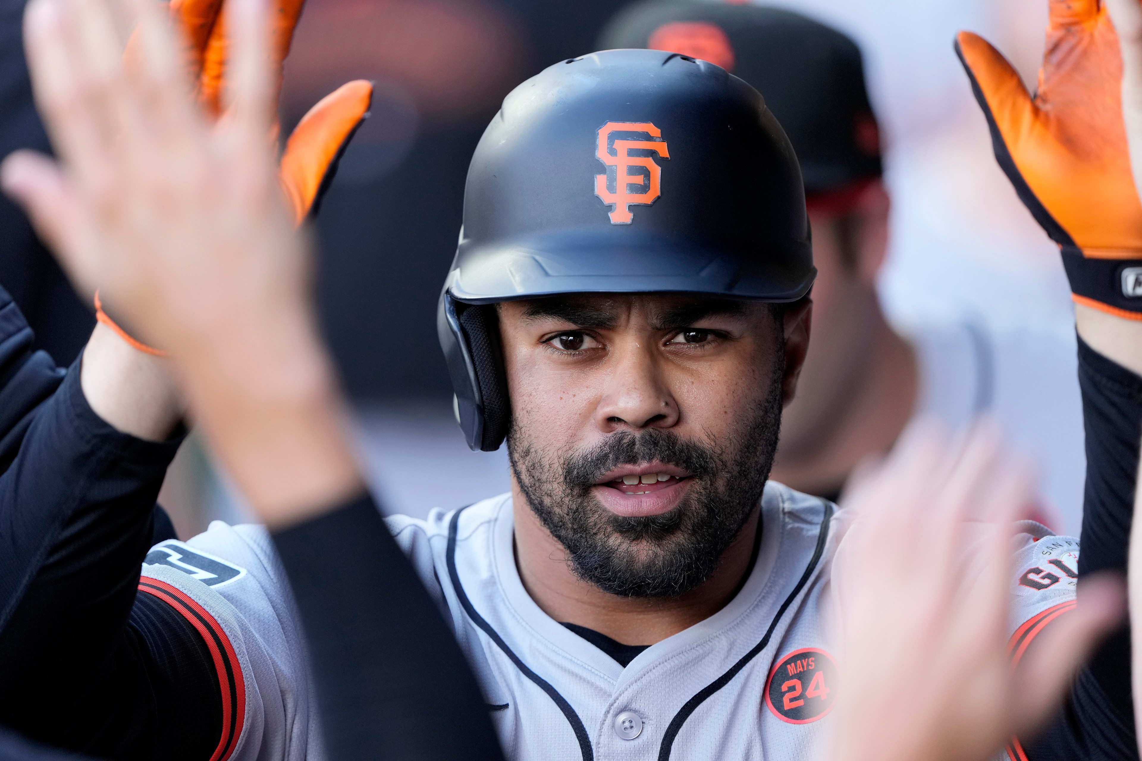 A baseball player wearing a black helmet with an orange logo is giving high-fives while wearing orange gloves. He's in a gray jersey with button details.