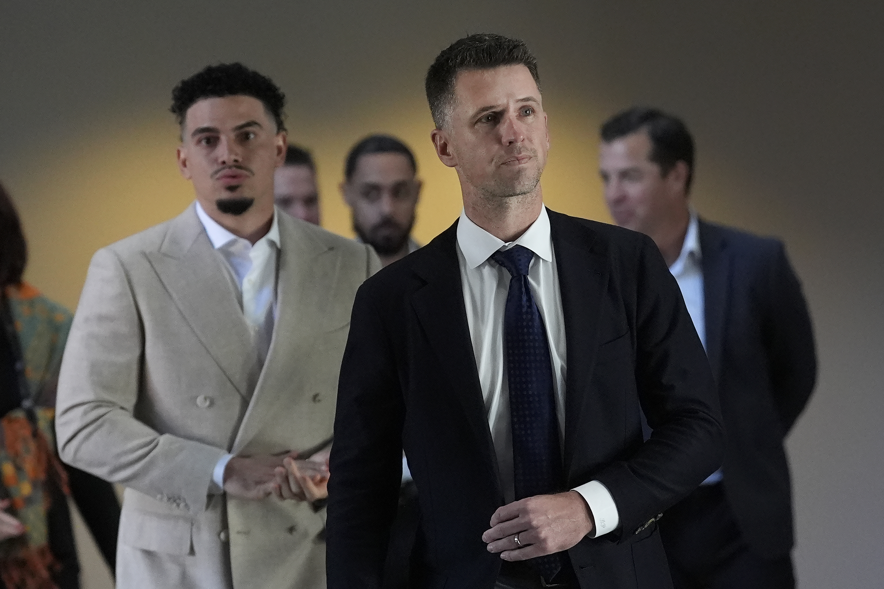 A group of men in suits stand together. The foreground shows two men, one in a light suit, the other in a dark suit and tie, both with serious expressions.