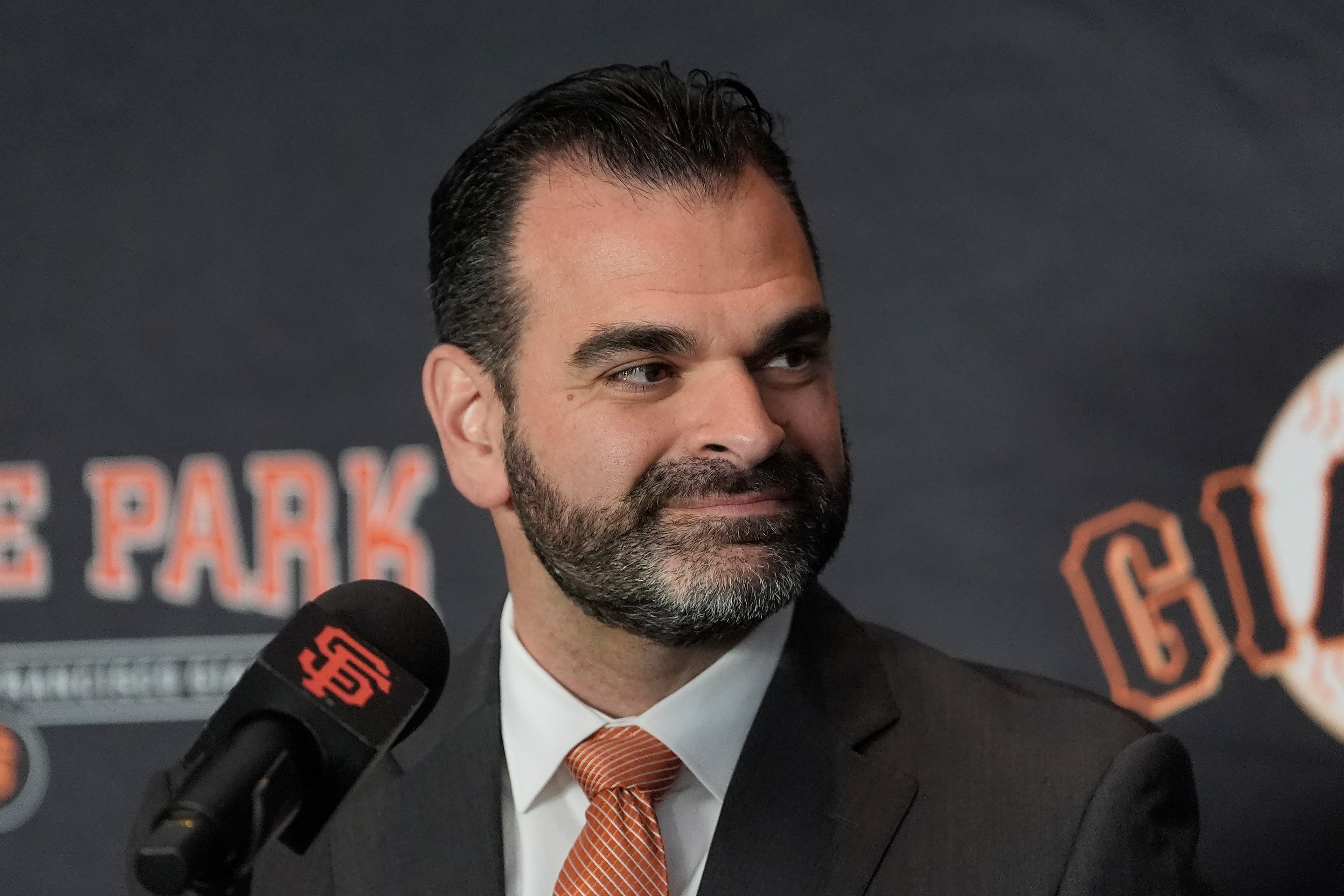 A man in a suit with a beard smiles slightly, standing in front of a backdrop with the words "GIANTS" and "PARK." A microphone is in front of him.