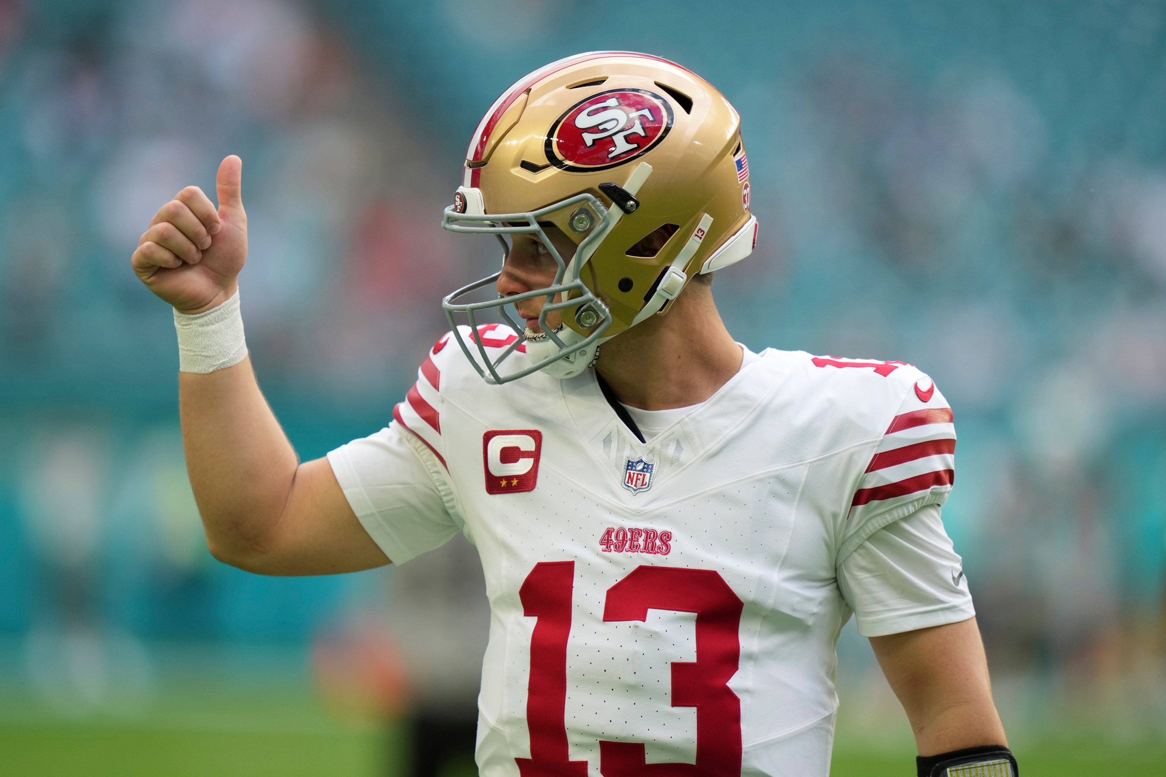 A football player wearing a San Francisco 49ers jersey and helmet gives a thumbs-up on a field, displaying jersey number 13. The background is a blurred crowd.