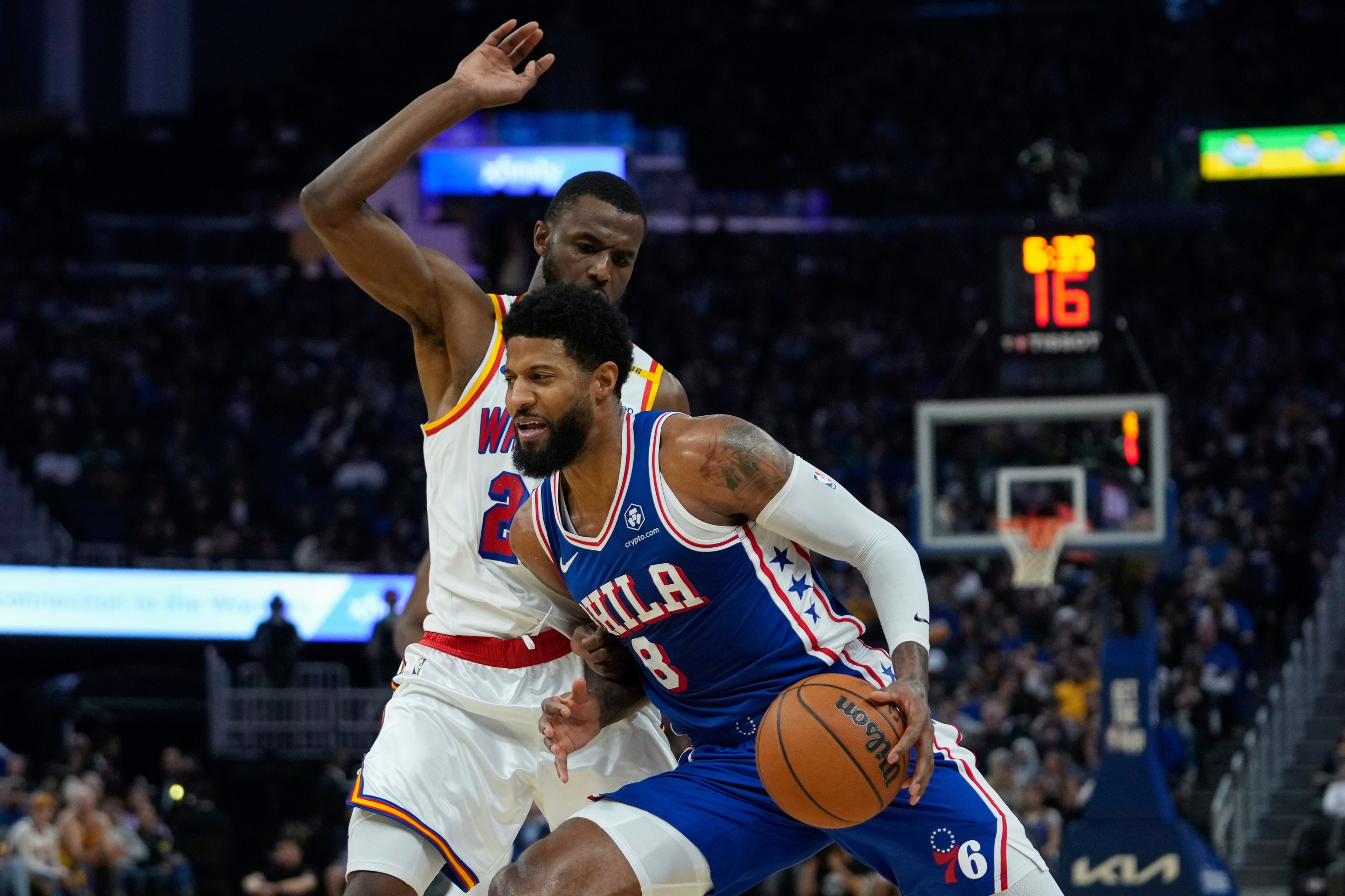 A basketball player in a blue &quot;Phila&quot; jersey drives past an opponent in a white &quot;W&quot; jersey. The scene is intense with a crowd watching and a scoreboard visible.
