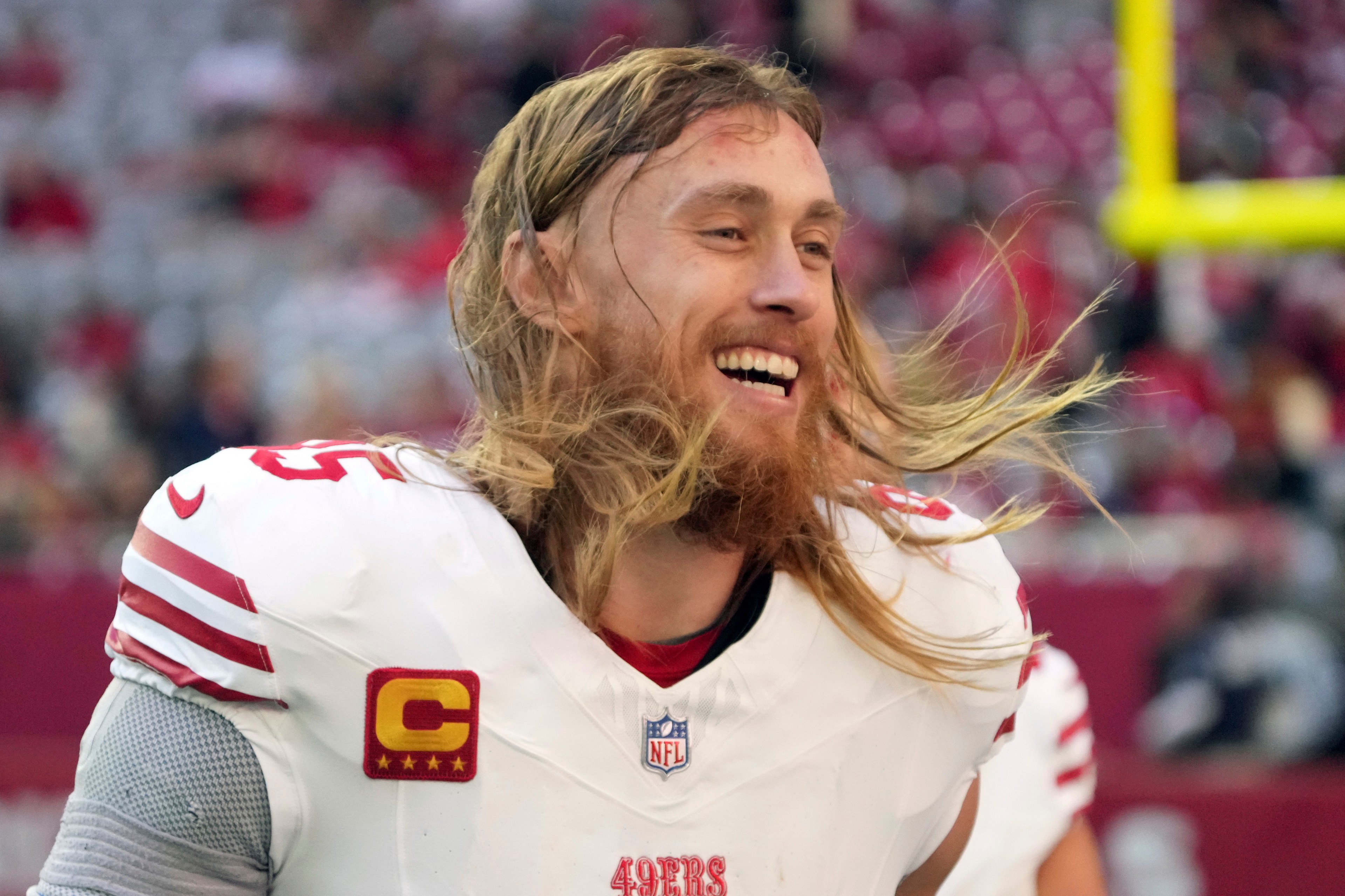 A smiling football player with long hair is wearing a white jersey with red stripes, featuring an NFL and captain's patch, in a stadium with a blurred background.