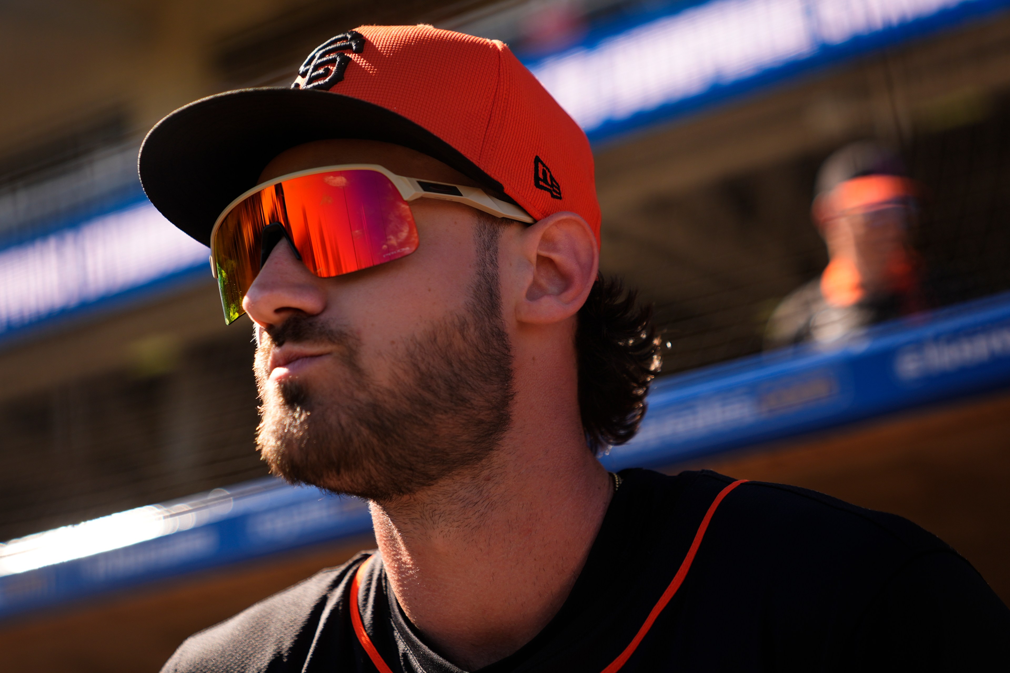 A person wearing an orange baseball cap and reflective sunglasses looks to the side. They have facial hair and wear a black sports shirt with orange trim.