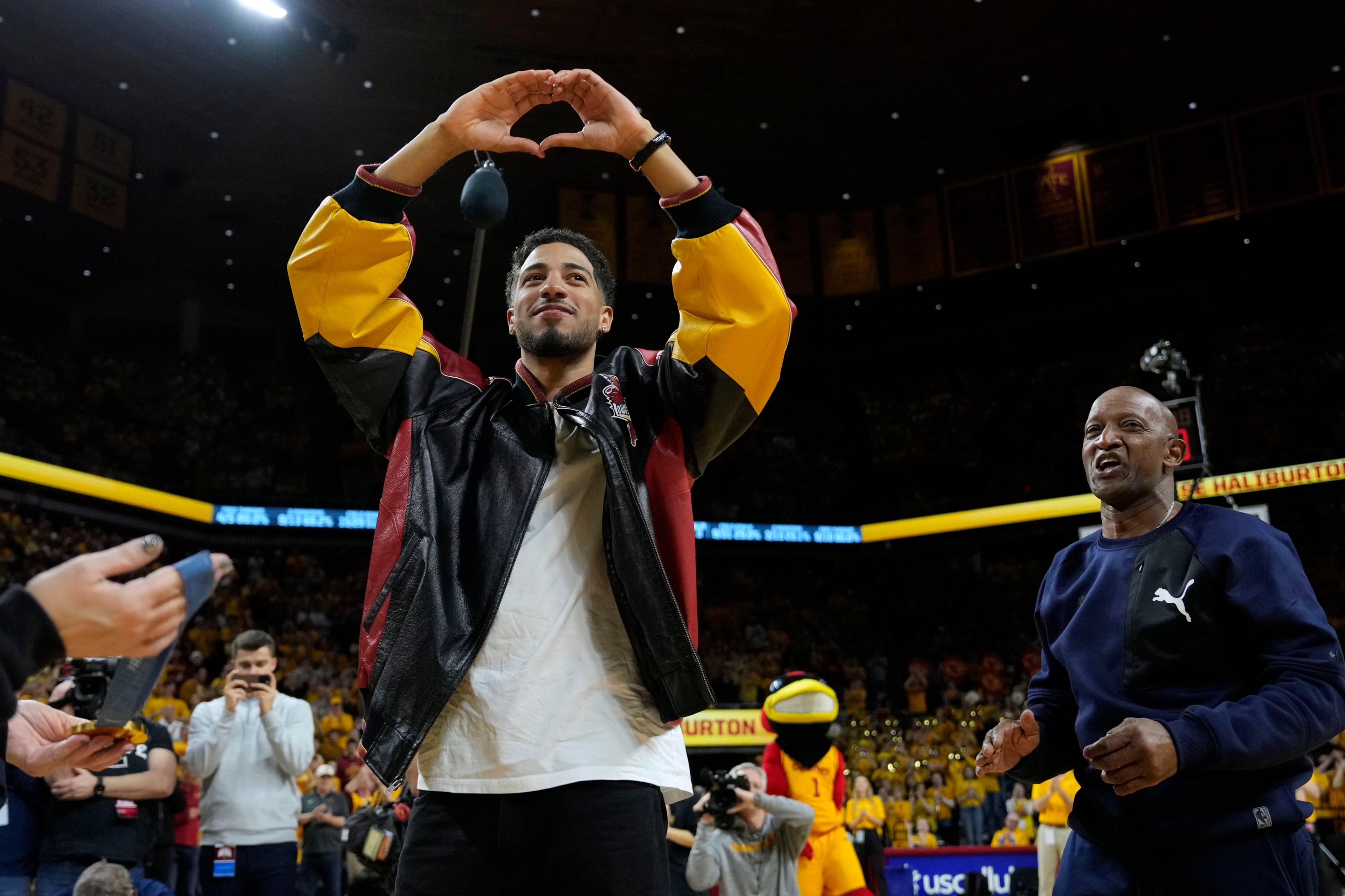 A person in a sports arena, wearing a colorful jacket, makes a heart shape with their hands. A man in a tracksuit stands nearby, and a mascot is visible behind them.