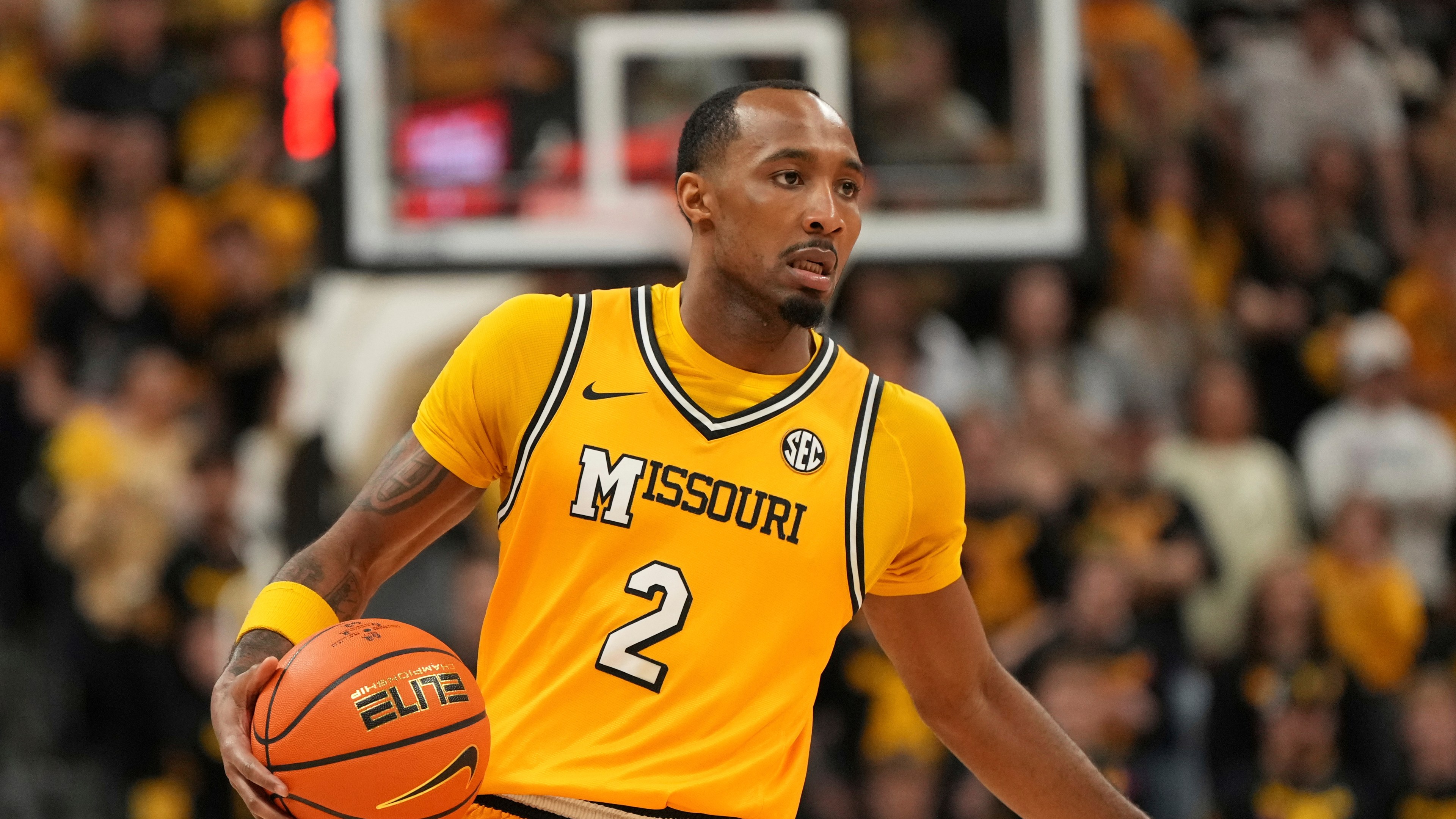A basketball player in a yellow &quot;Missouri&quot; jersey with the number 2 is dribbling the ball on a court, amidst a blurred crowd background.