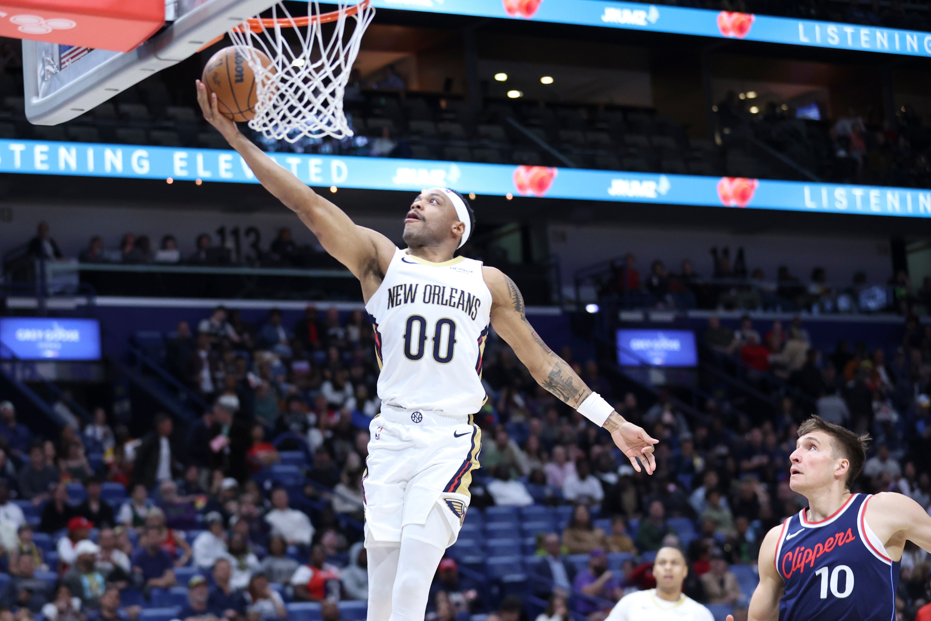A basketball player from the New Orleans team is doing a layup while an opposing player from the Clippers watches. The crowd is visible in the background.