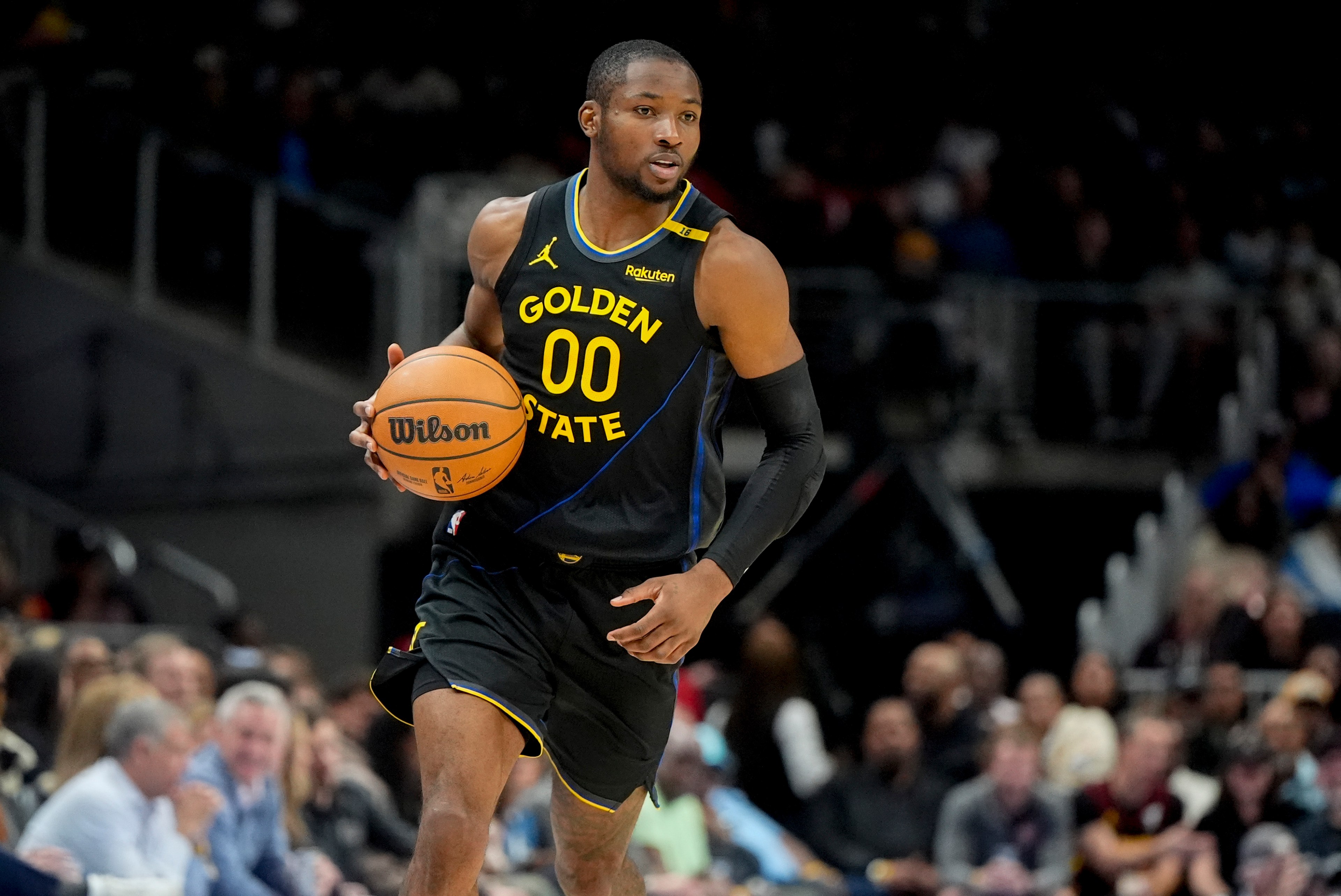 A basketball player wearing a black Golden State jersey dribbles a ball down the court. The audience is blurred in the background.