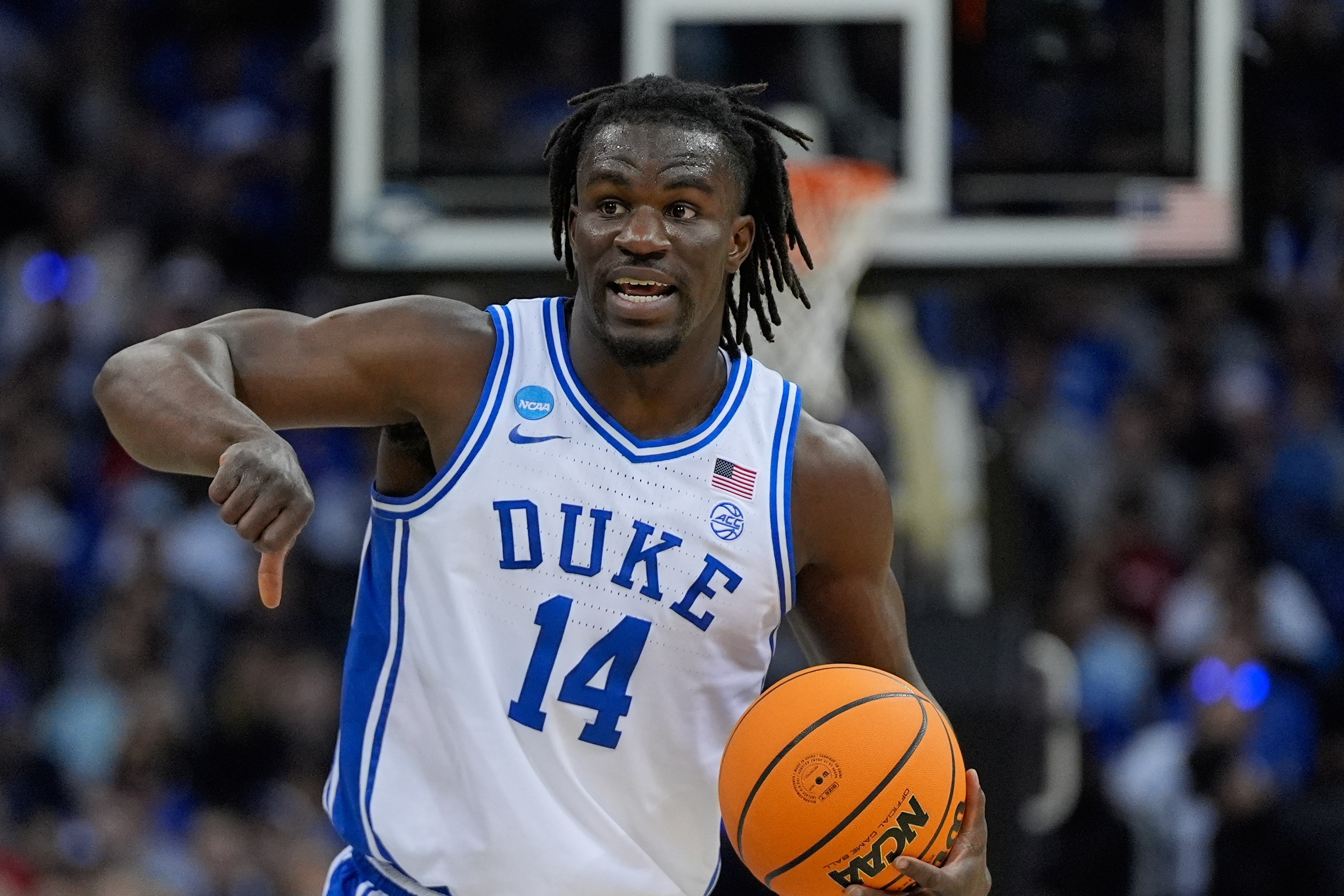 A basketball player wearing a Duke jersey with the number 14 is dribbling a ball on the court. The background shows a blurred crowd.