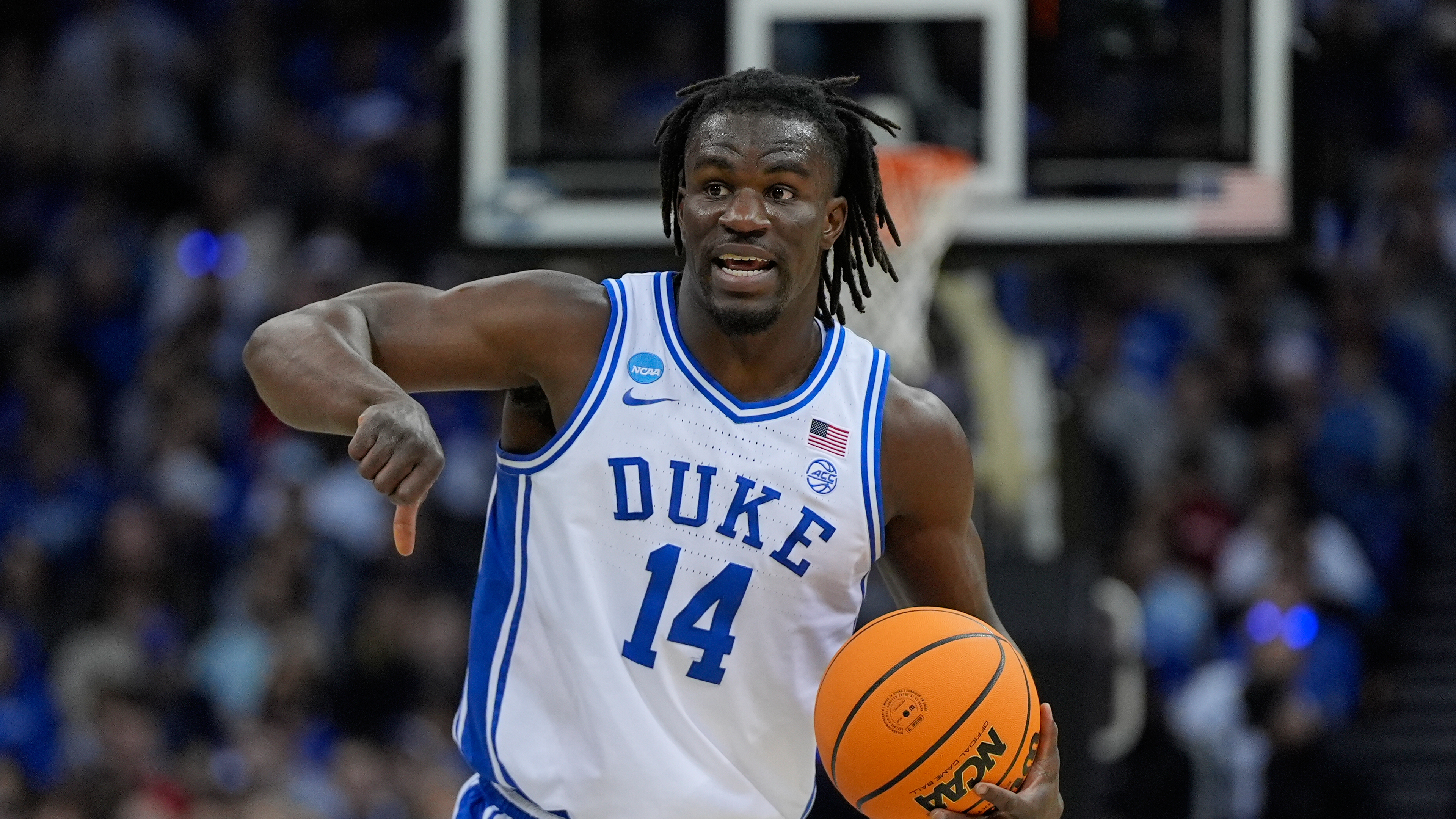 A basketball player wearing a Duke jersey with the number 14 is dribbling a ball on the court. The background shows a blurred crowd.