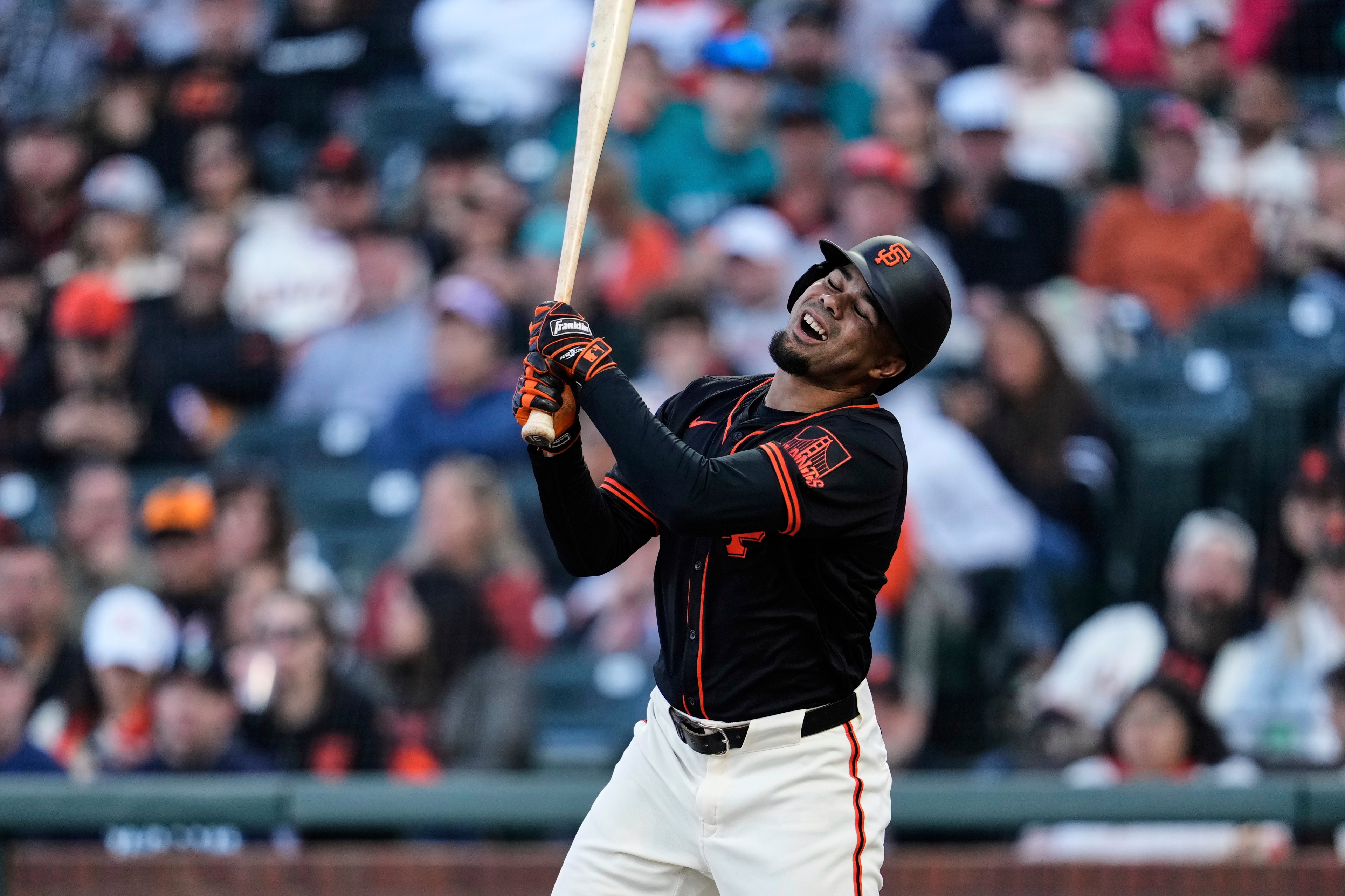 A baseball player in a black and orange uniform is mid-swing with a pained expression. He wears a helmet and gloves, and the crowd is blurred in the background.