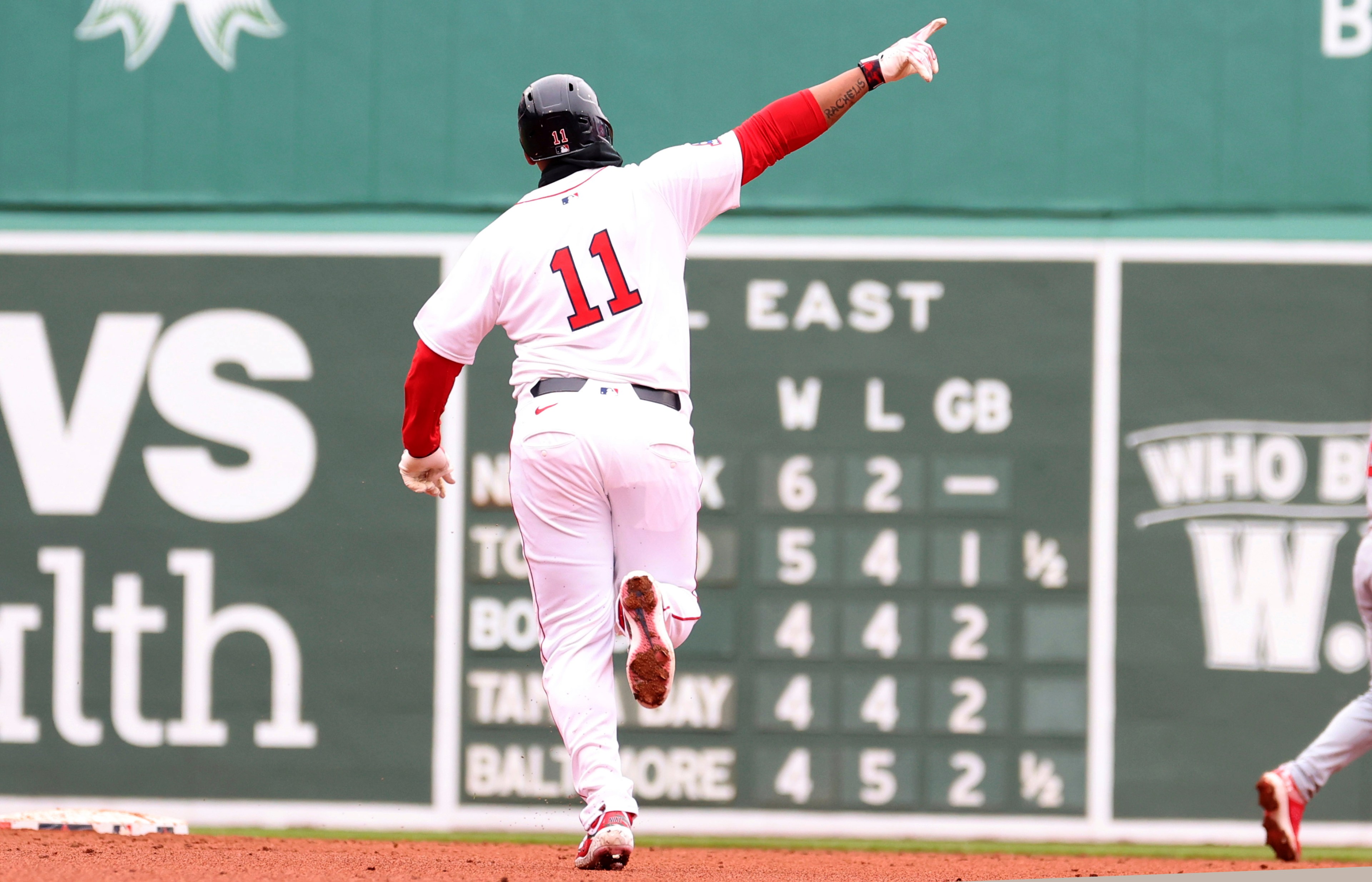 A baseball player in a white uniform with number 11 runs on a field, pointing upwards. Behind is a green scoreboard displaying team standings.